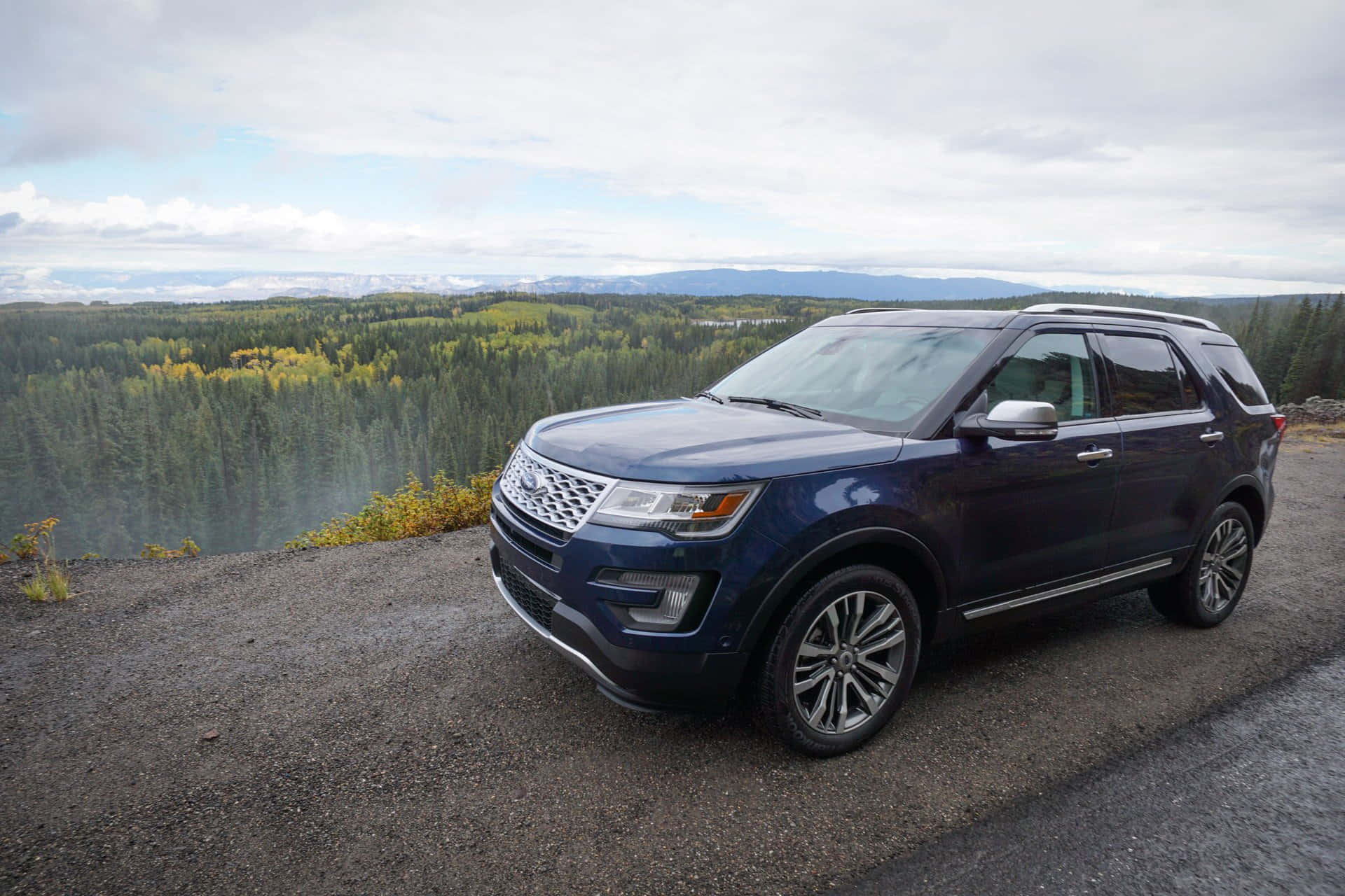 Ford Explorer 桌布 壁紙 — 一輛深藍色福特Explorer SUV停泊在一個碎石觀景點，俯瞰著廣闊的森林山谷和遠處的山脈，天空多雲。