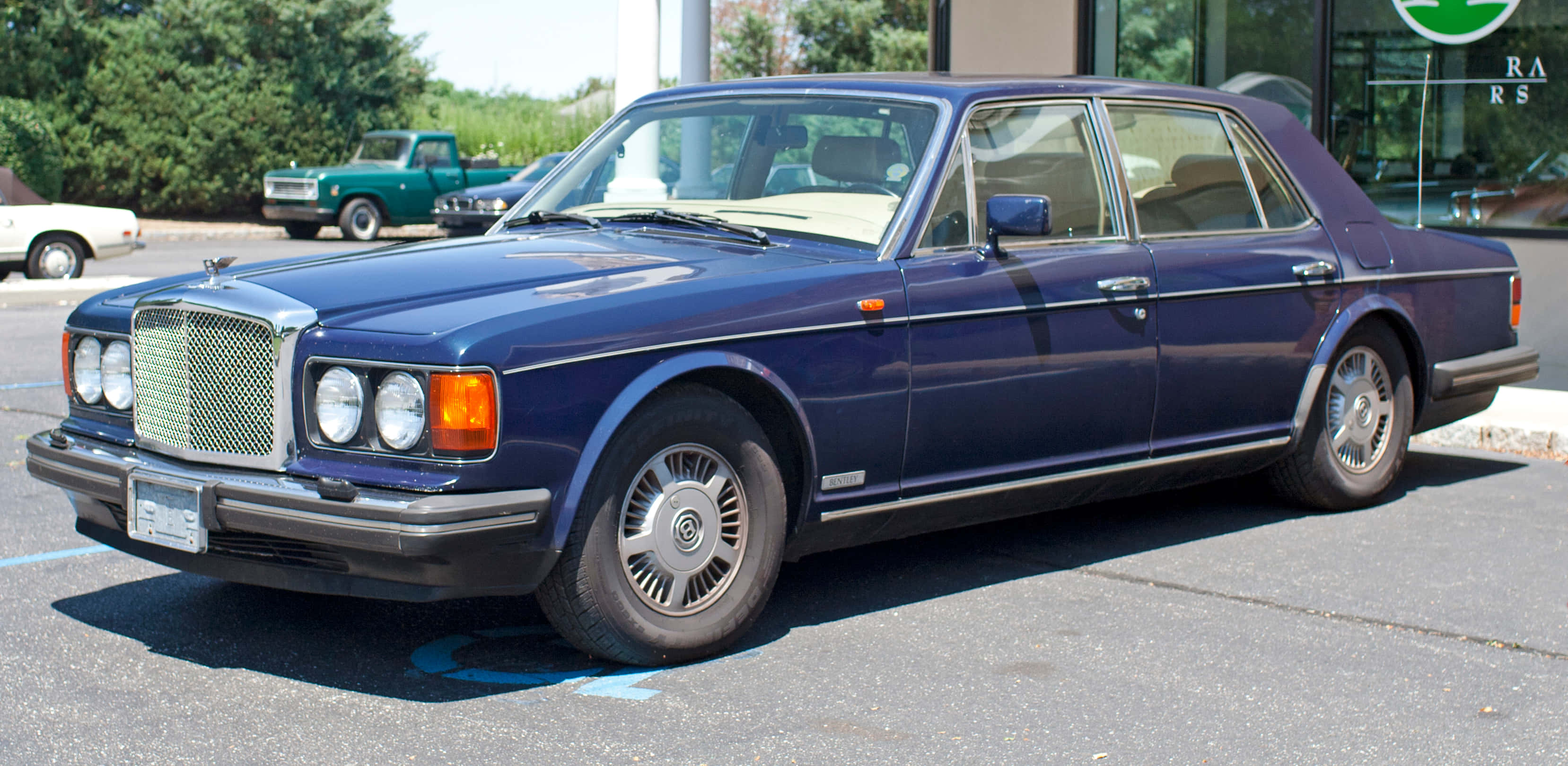 Bentley Eight 桌布 壁紙 — 一輛深藍色賓利轎車在明亮陽光下閃閃發光，停放在一個附近有其他復古汽車的停車場。