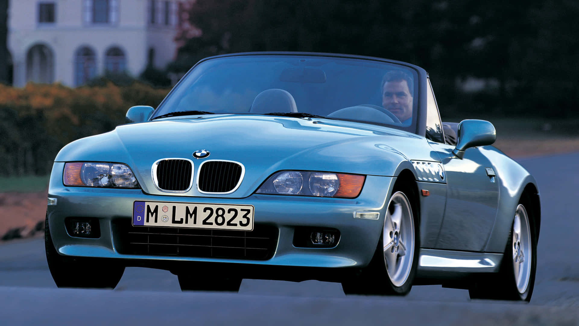 Bmw Z3 桌布 壁紙 — 一輛淺藍色BMW Z3跑車，司機微笑著，在一條大宅附近的林蔭大道上行駛。