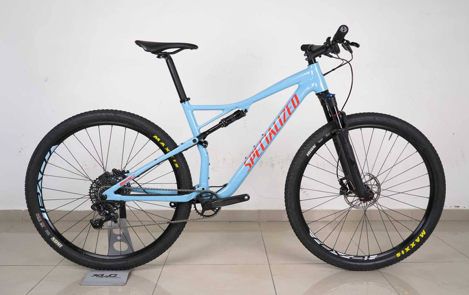 腳踏車 桌布 壁紙 — 一輛淺藍色Specialized登山自行車，配備黑色輪胎和紅色品牌標誌，直立於磁磚地板上，背景為純白牆壁。