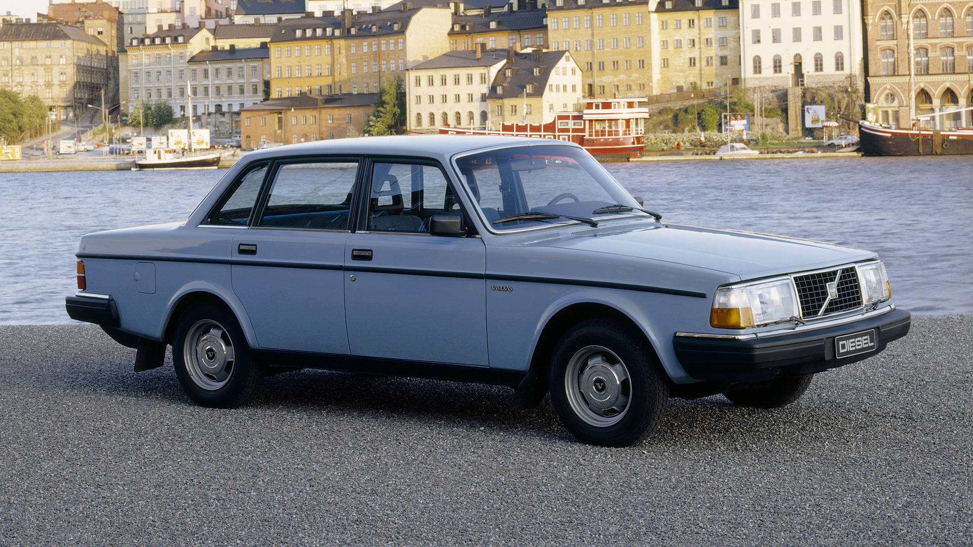 volvo iPhone 桌布 壁紙 — 一輛淺藍色富豪240柴油轎車停在海濱的碎石路上，背景有歷史建築和船隻。