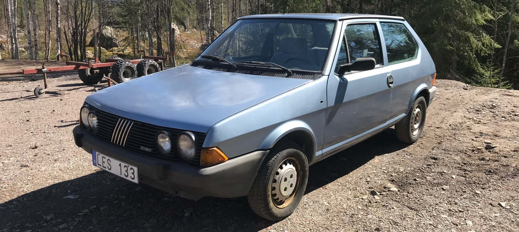 Fiat Ritmo 桌布 壁紙 — 一輛淺藍色復古飛雅特Uno停放在碎石地上，周圍是高聳的松樹，在明亮的日光下。