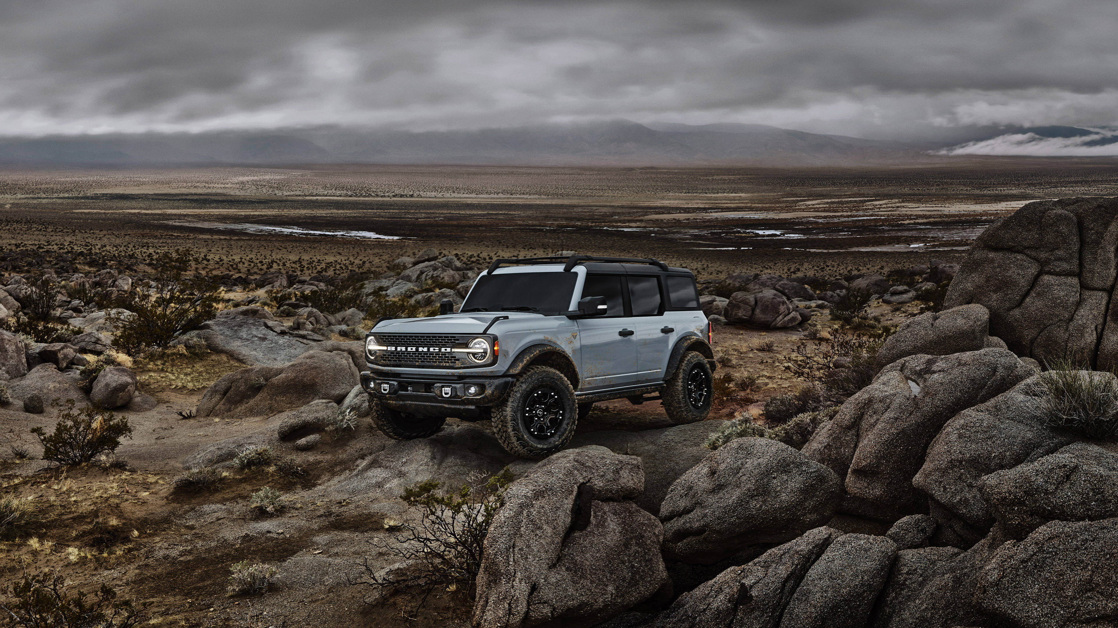 Ford Bronco 桌布 壁紙 — 一輛淺藍色的福特 Bronco 堅固地矗立在岩石地形上，背景是陰沉的陰天和遠處的山脈。