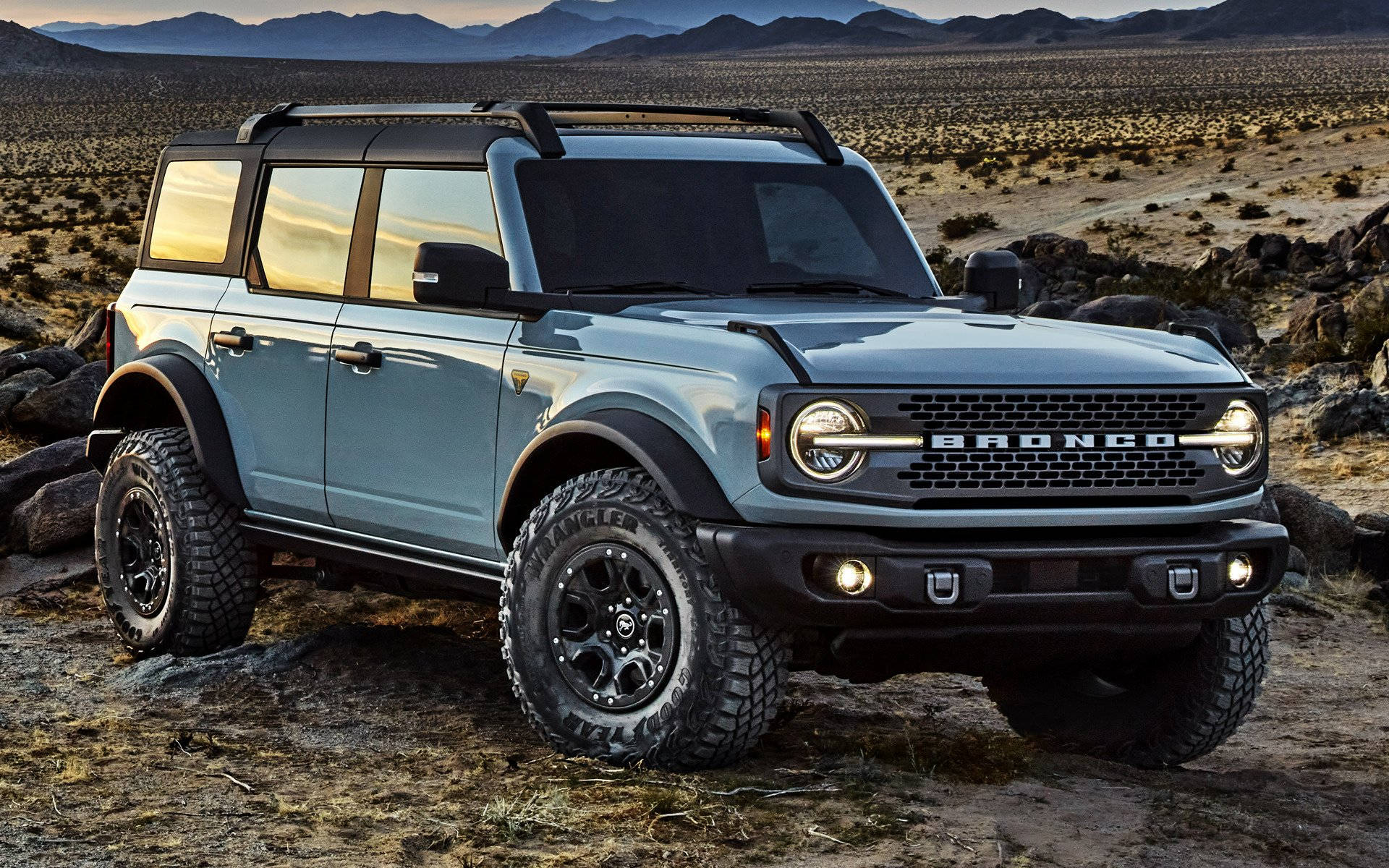 Ford Bronco 桌布 壁紙 — 一輛淺藍色福特Bronco SUV，帶有黑色飾邊和堅固輪胎，在金色日落天空和遠山下的岩石沙漠地形上蓄勢待發。