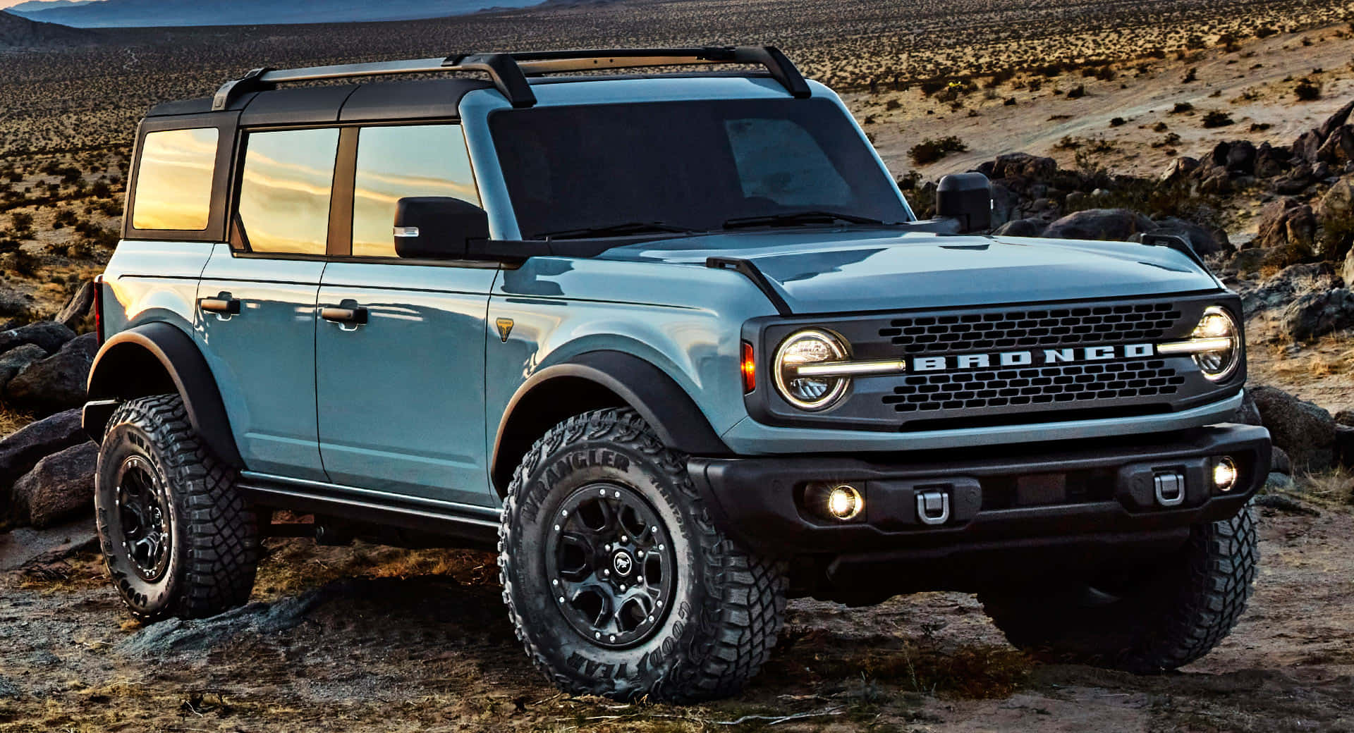 Ford Bronco 桌布 壁紙 — 一輛淺藍色福特Bronco SUV，配備黑色飾邊和堅固輪胎，在溫暖的日落餘暉下，傲然屹立在岩石沙漠地形上。