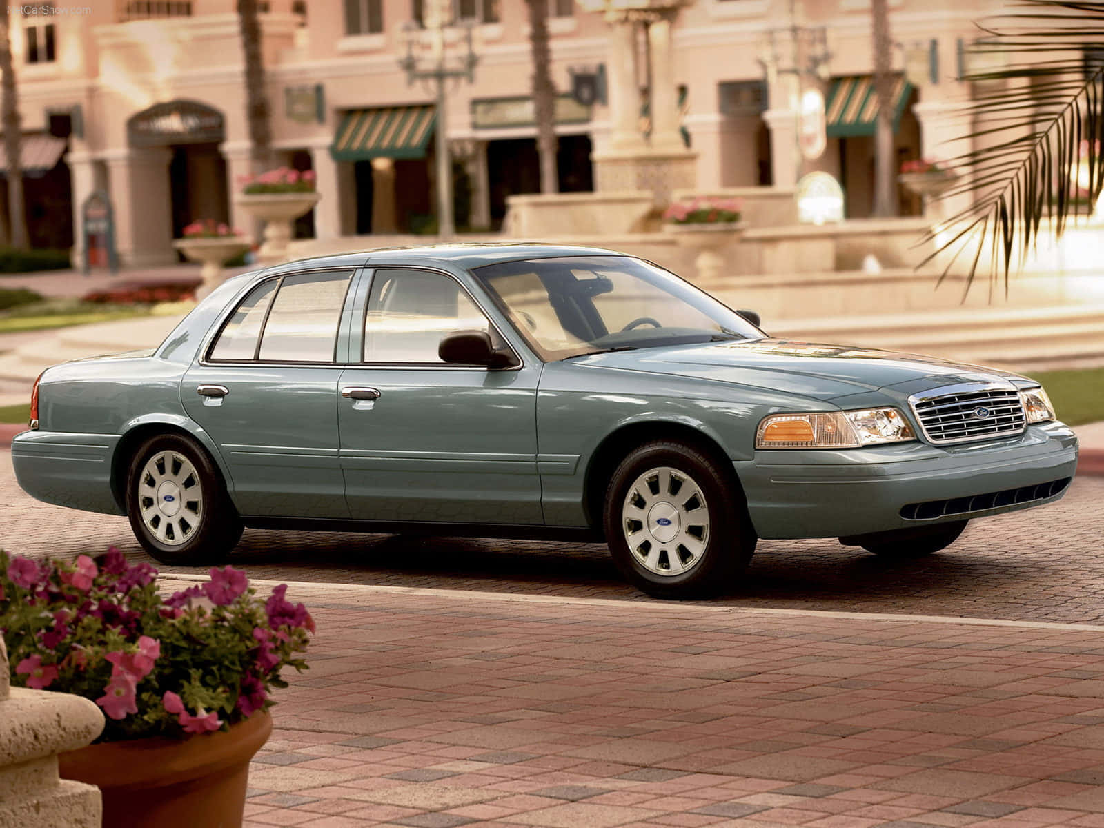 Ford Crown Victoria 桌布 壁紙 — 一輛淺藍色福特皇冠維多利亞轎車停放在磚砌廣場上，周圍有棕櫚樹和商店門面，沐浴在溫暖的黃金時刻光線中。
