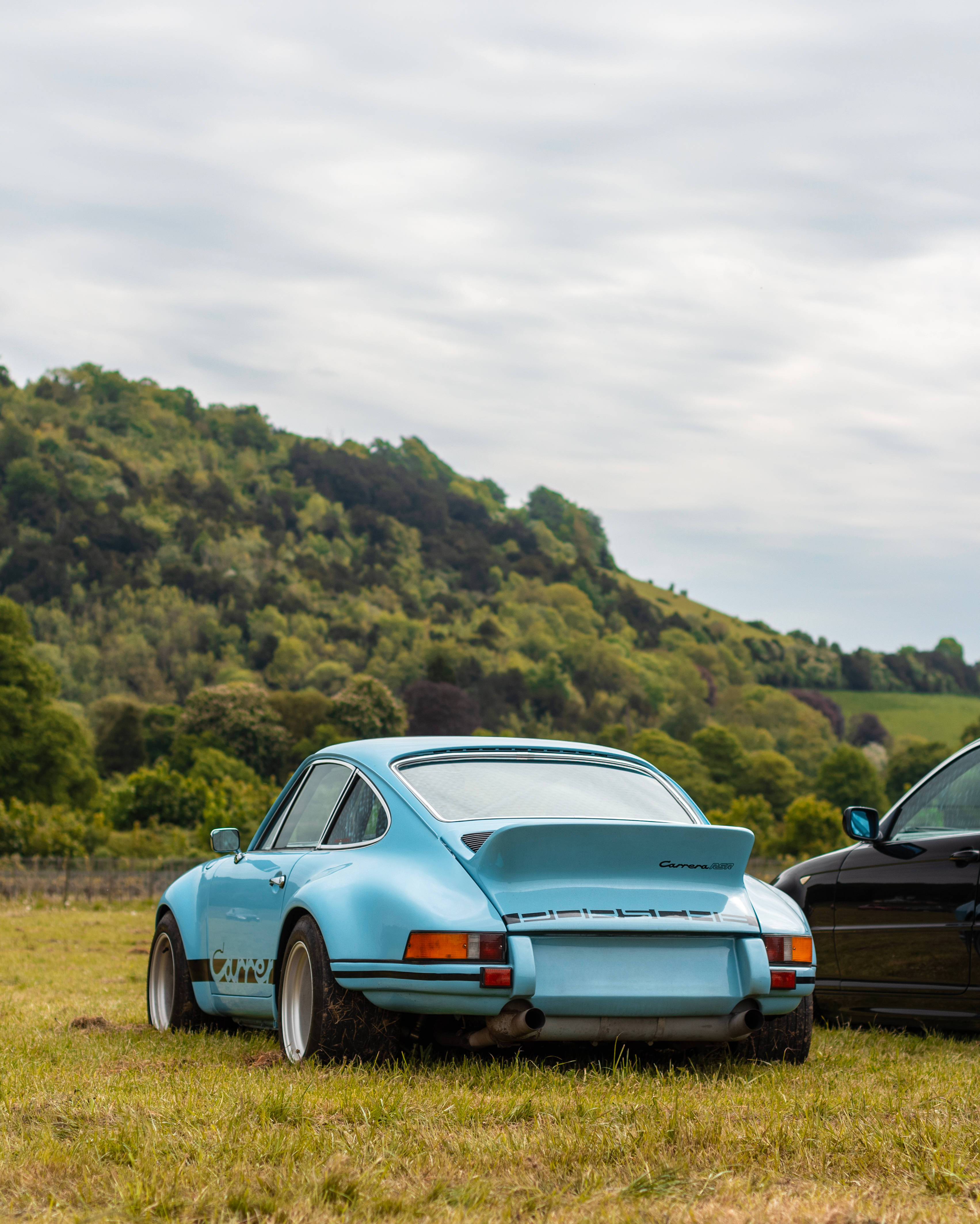 Luxury 桌布 壁紙 — 一輛淺藍色經典保時捷911 Carrera RS停放在草地上，背景是森林覆蓋的山丘，天空多雲。