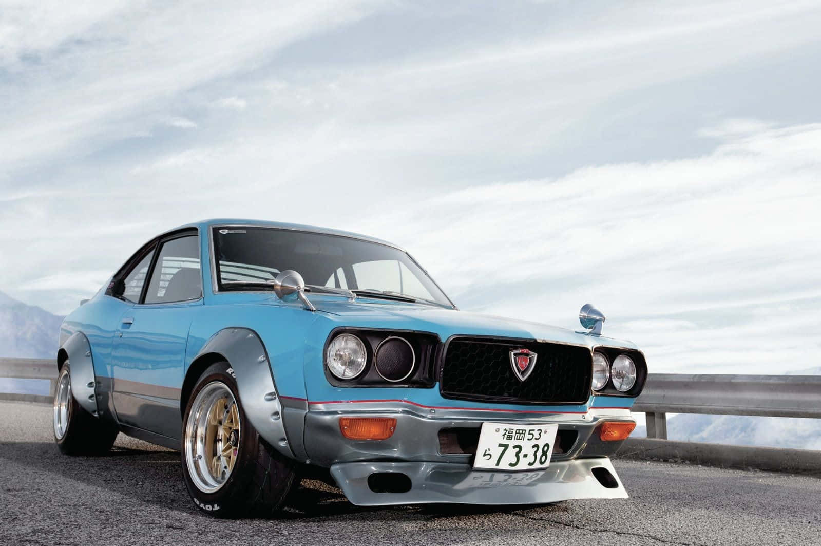 Mazda Rx-3 桌布 壁紙 — 一輛淺藍色經典日本跑車，配備寬體套件和金色輪圈，停泊在陰天天空下的山路上。