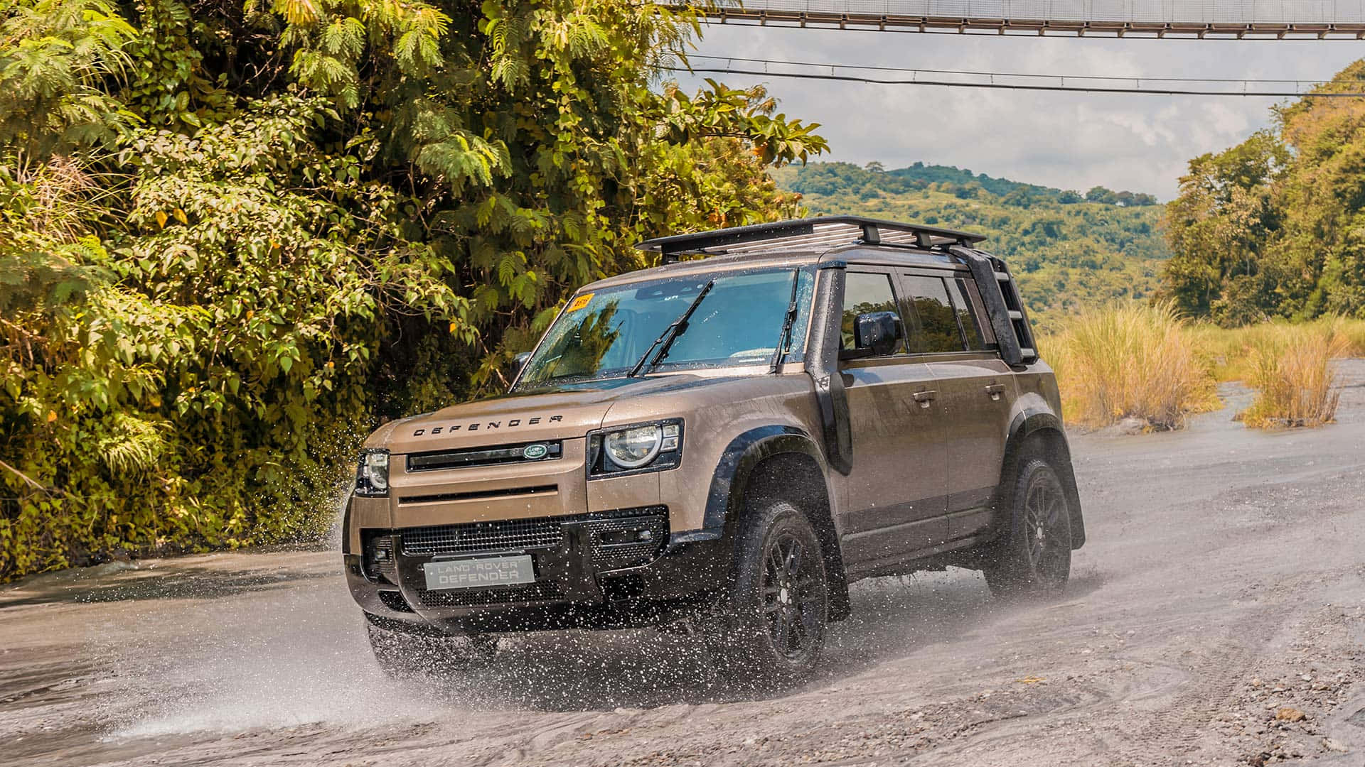 Land Rover Defender 桌布 壁紙 — 一輛淺褐色 Land Rover Defender 在陰天下的森林泥路上濺起泥水。