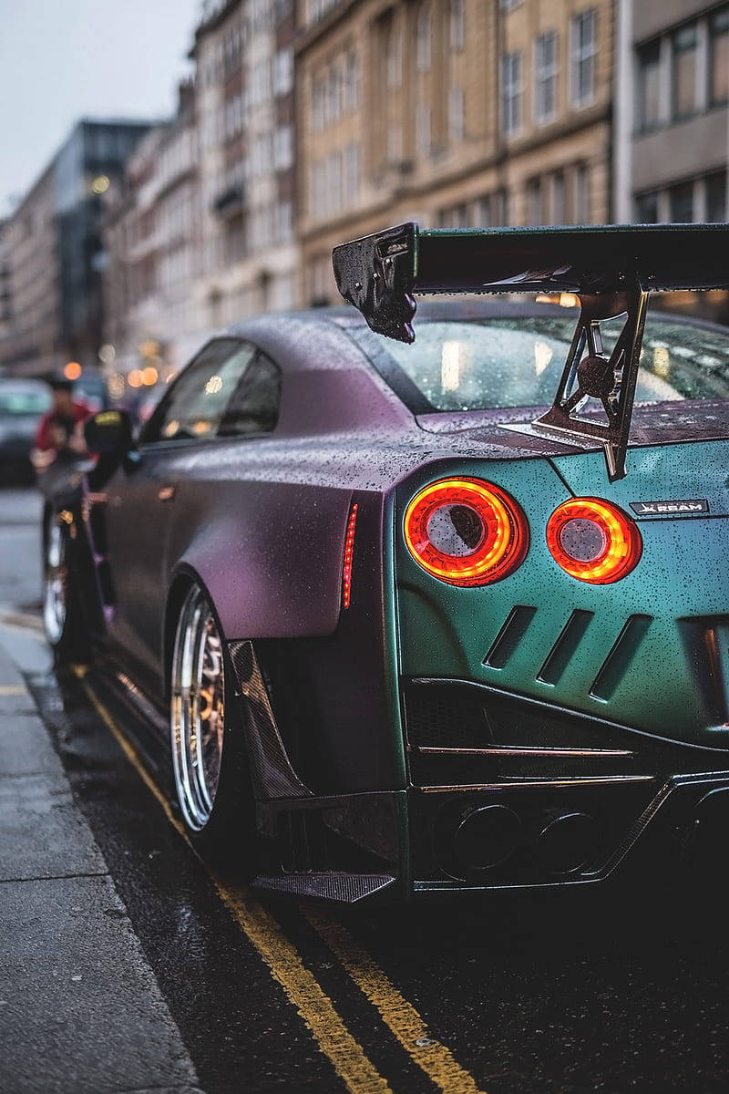 Best Car 桌布 壁紙 — 一輛濕透的紫色和青色日產GT-R，亮著紅色尾燈，停在雨中的城市街道上。