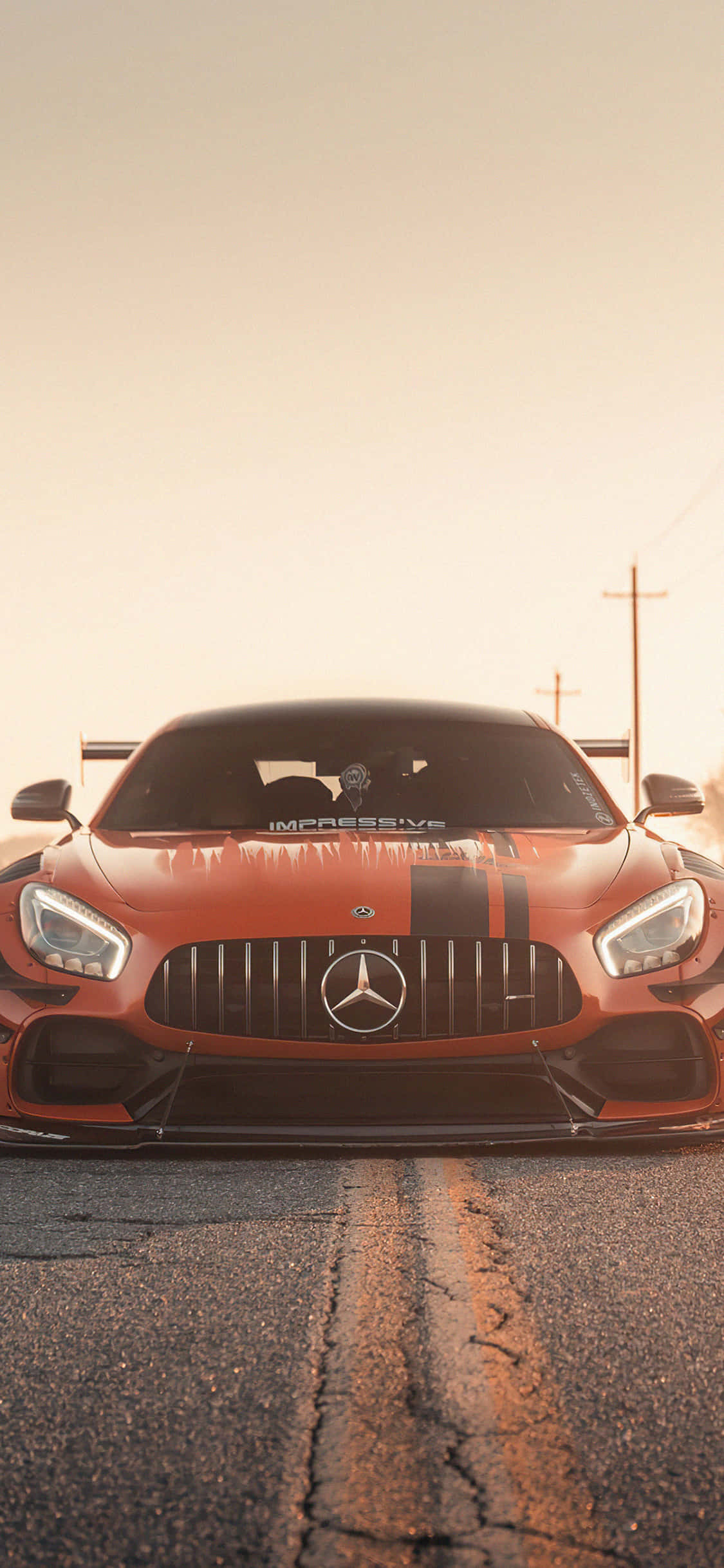 iPhone X Amg Gt-r 桌布 壁紙 — 一輛火橙色Mercedes-AMG GT賽車在龜裂的柏油路上向前行駛，背景是朦朧的金色日落。