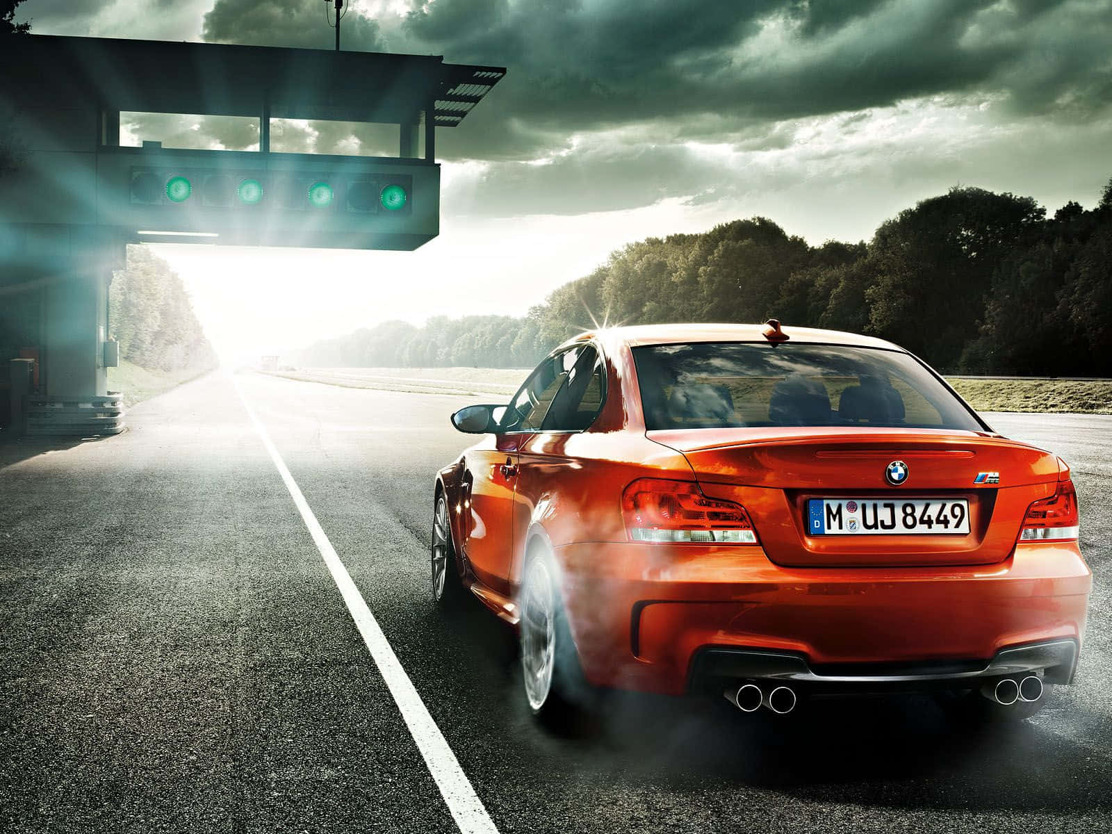 Bmw 1 Series 桌布 壁紙 — 一輛火橙色的BMW M1跑車從賽車場隧道中轟鳴而出，輪胎冒著濃煙，背景是戲劇性的多雲天空。