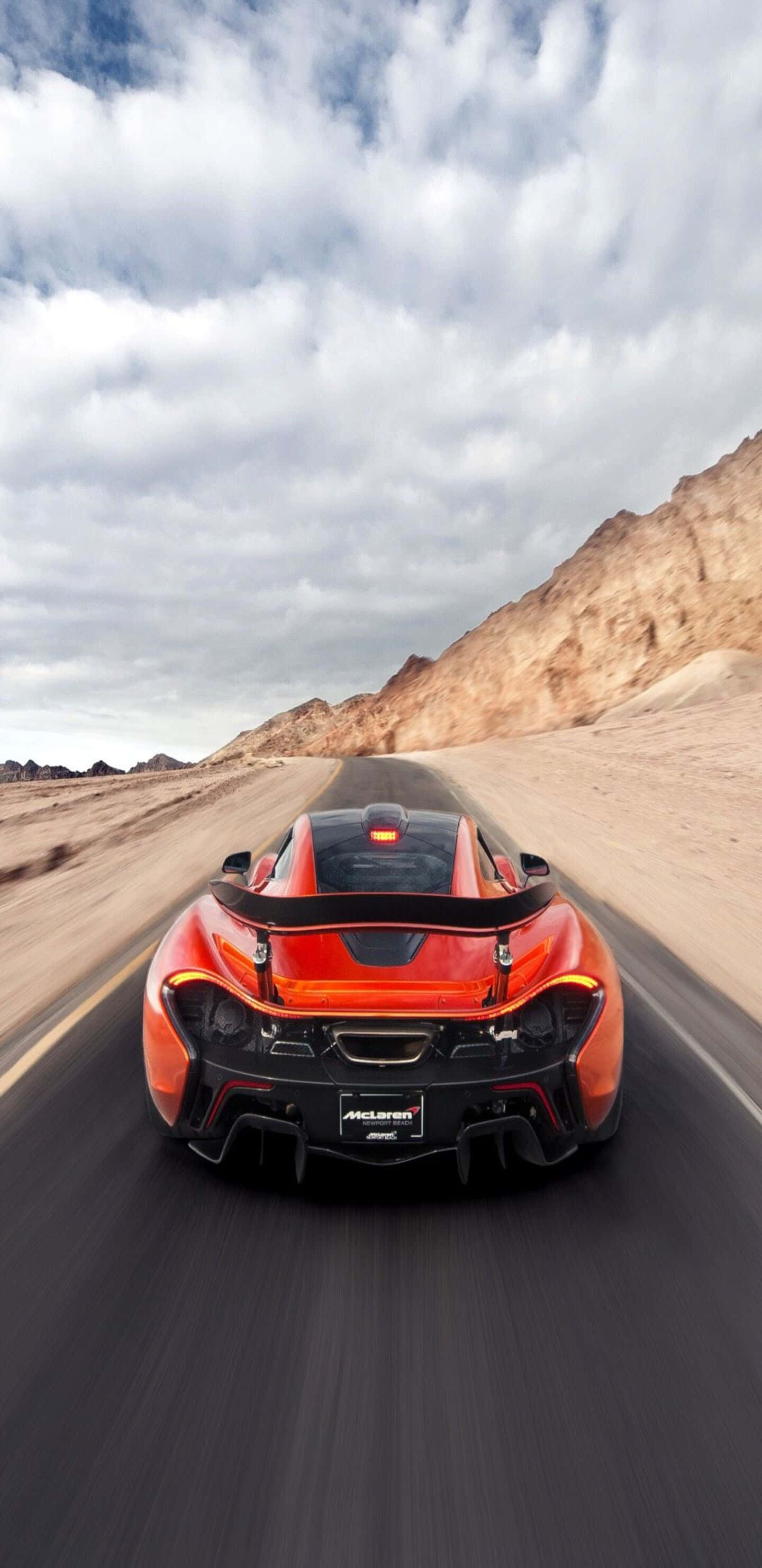 人像動態 桌布 壁紙 — 一輛火橙色的McLaren跑車在雲層密布的天空下，沿著一條有岩石山丘的沙漠公路飛馳。