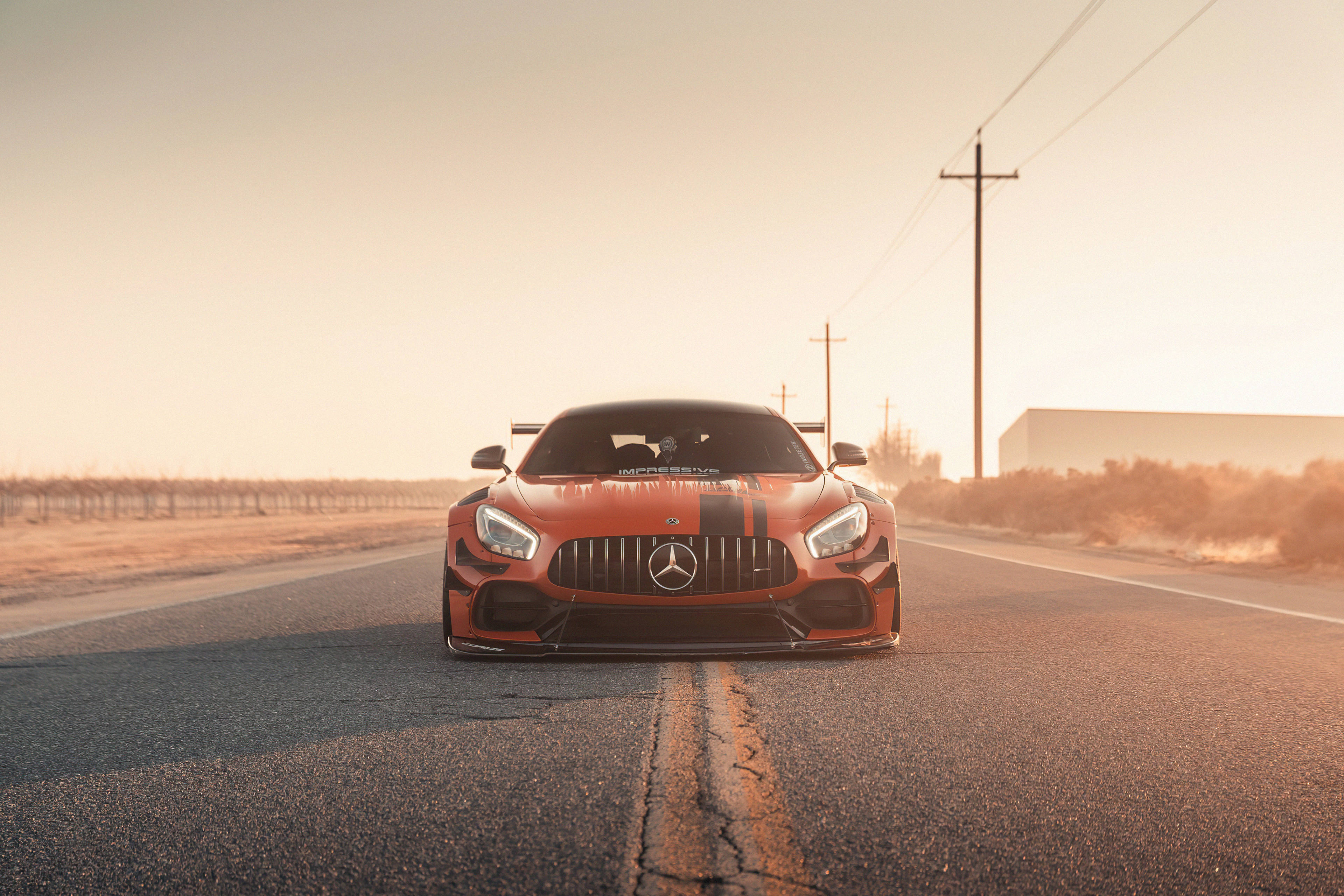 4K 奔馳壁紙 桌布 壁紙 — 一輛火橙色的Mercedes-AMG GT R在黃金時段面向前方，行駛在裂開的瀝青路上，背景有電線和乾燥的田野。