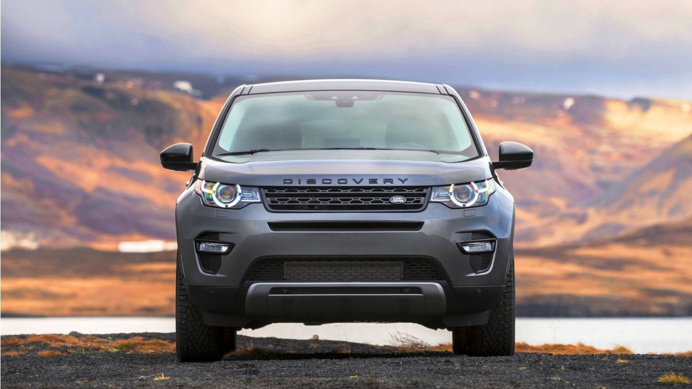 沙漠 桌布 壁紙 — 一輛灰色 Land Rover Discovery SUV 在礫石灘上向前行駛，背景是金色山丘和寧靜的湖泊，天空呈現柔和的暮色。
