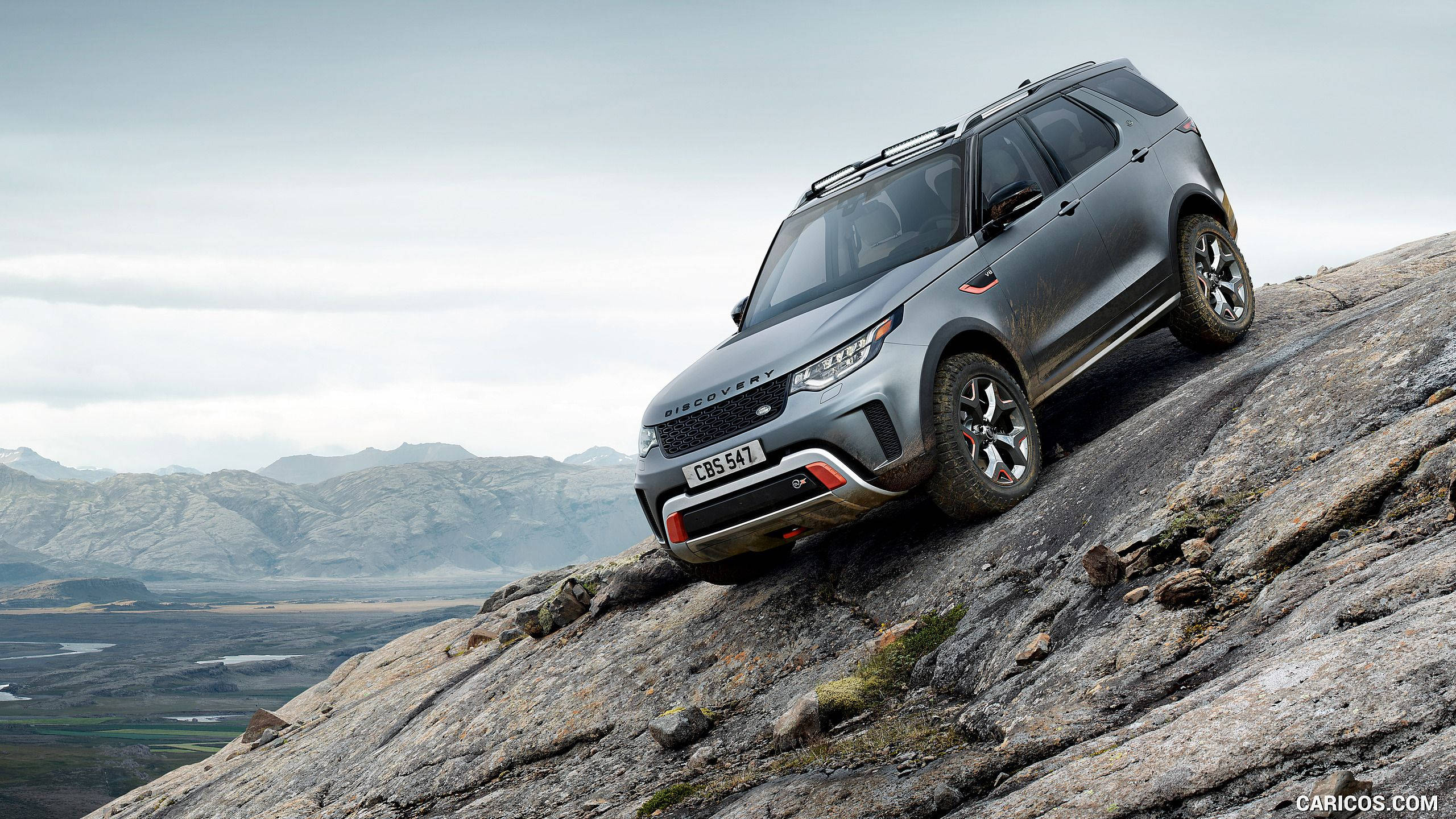 荒原路華 桌布 壁紙 — 一輛灰色 Land Rover Discovery 征服陡峭岩石斜坡，山脈和山谷在陰天天空下延伸。