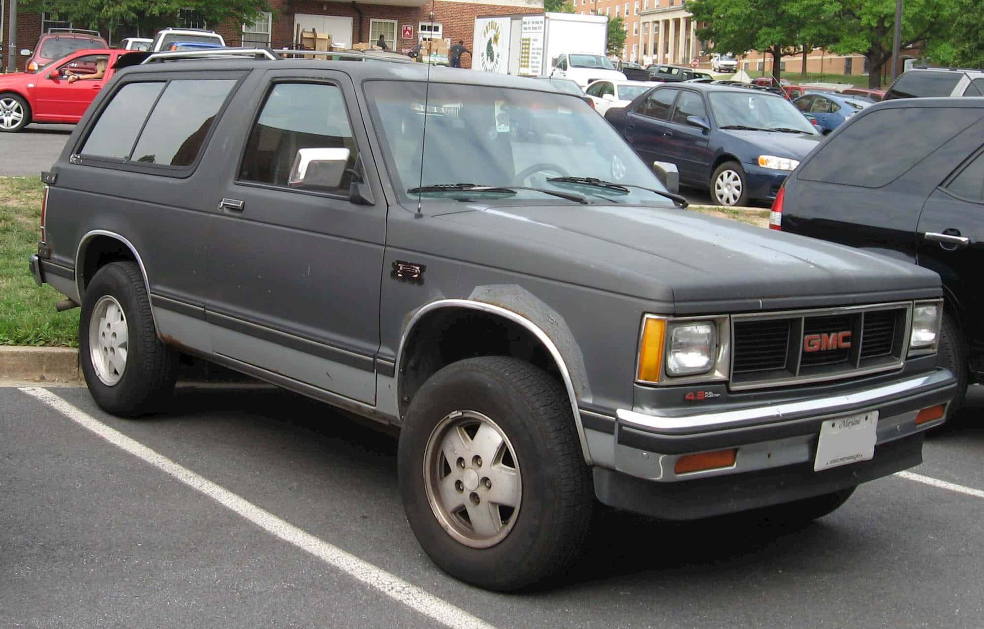 Gmc Jimmy 桌布 壁紙 — 一輛灰色1980年代GMC Safari SUV停放在日間停車場，背景有其他汽車和磚砌建築。