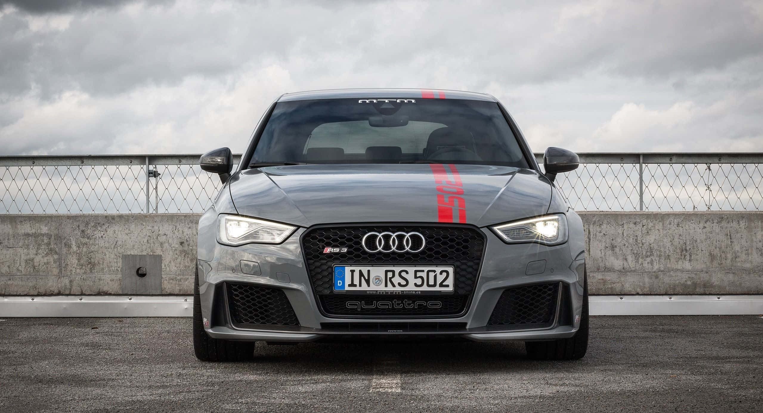 Audi Rs3 桌布 壁紙 — 一輛灰色Audi RS3帶有紅色賽車條紋，在陰天下的混凝土賽道上蓄勢待發。