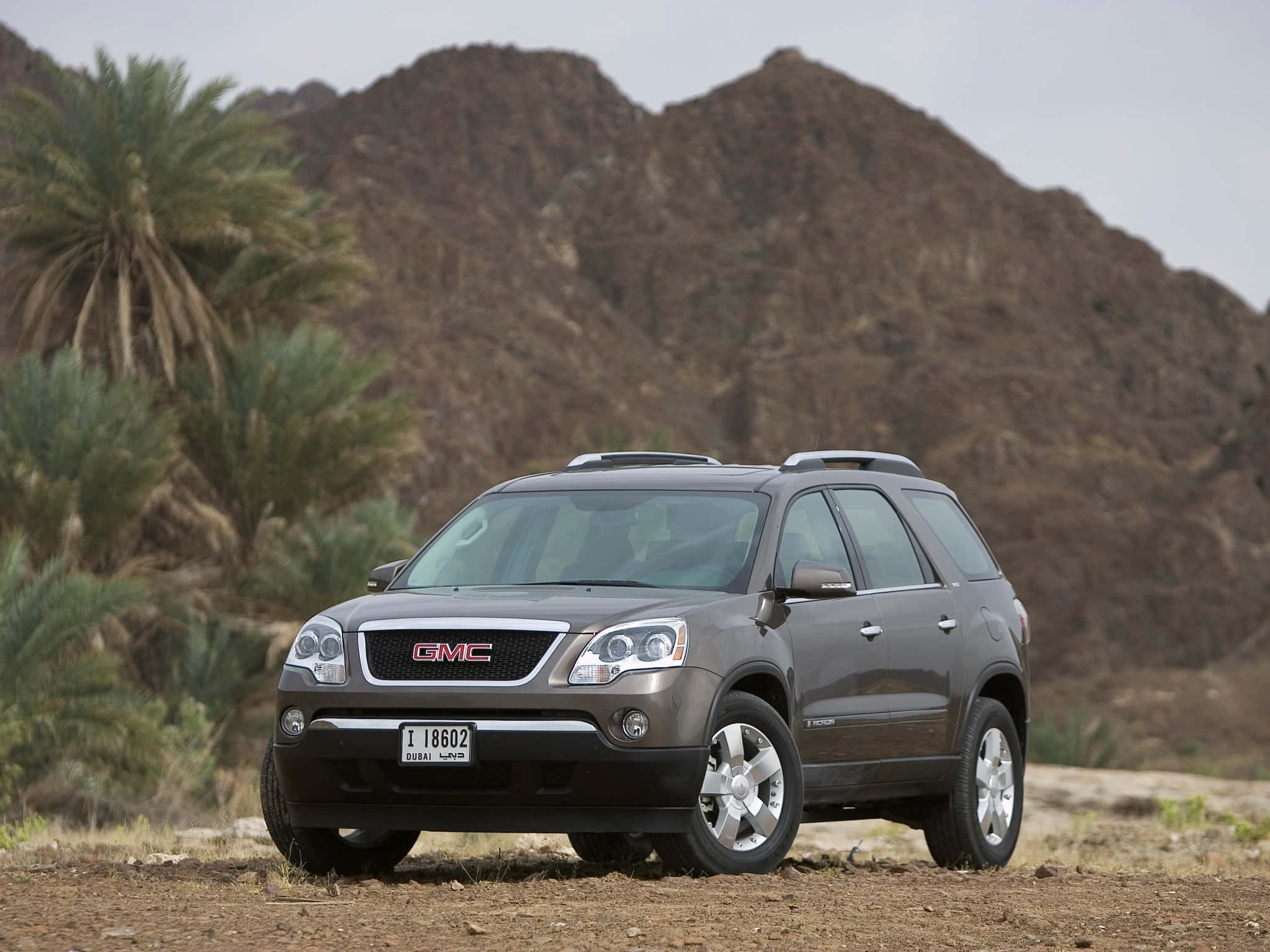 Gmc Acadia 桌布 壁紙 — 一輛灰色GMC Acadia SUV停泊在乾燥土地上，背景有棕櫚樹和岩石山丘，天空多雲。