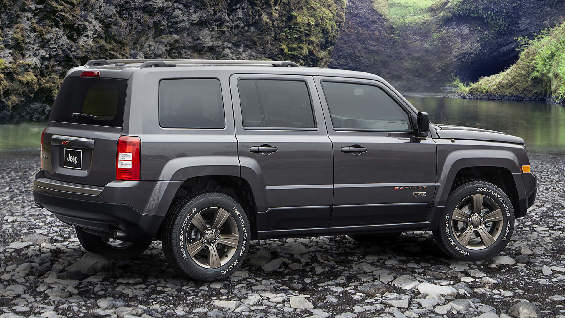Jeep Patriot 桌布 壁紙 — 一輛灰色Jeep Patriot SUV停泊在岩石河岸上，背後有長滿苔蘚的懸崖和寧靜的水面。