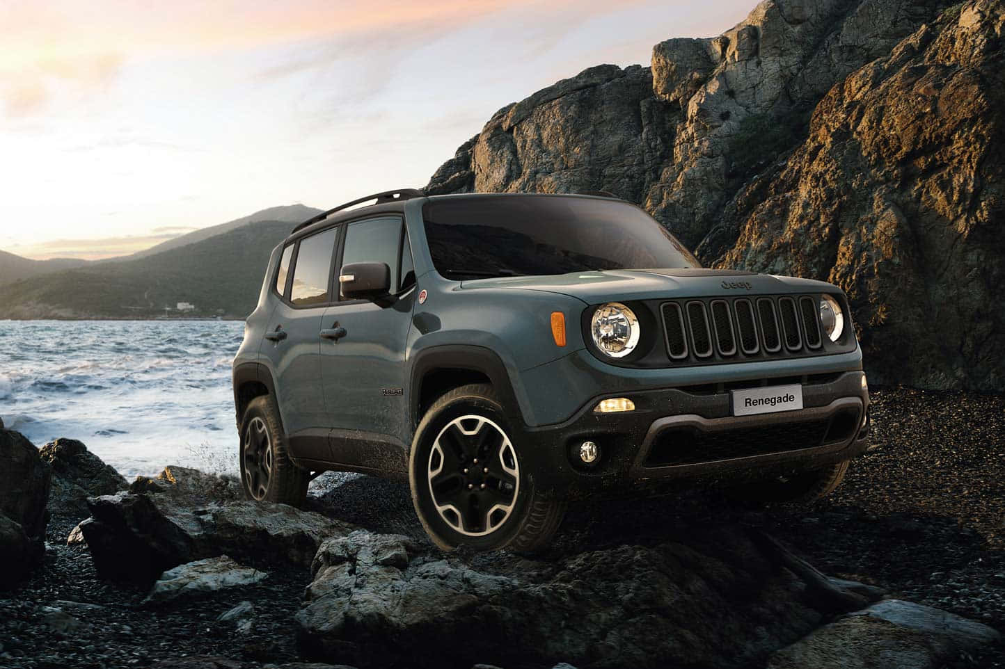 Jeep Renegade 桌布 壁紙 — 一輛灰色Jeep Renegade SUV停泊在岩石地形上，旁邊是日落時分波濤洶湧的海洋，背景有山脈。