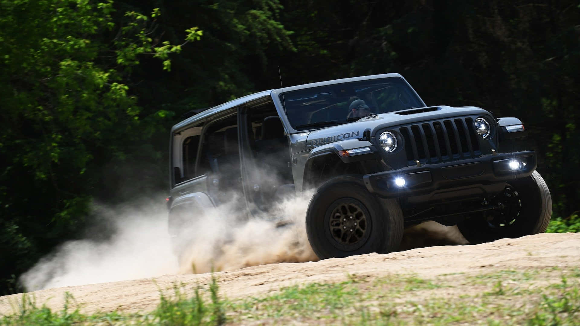 吉普 桌布 壁紙 — 一輛灰色Jeep Wrangler Rubicon在茂密翠綠的森林中行駛於泥土小徑，揚起灰塵。