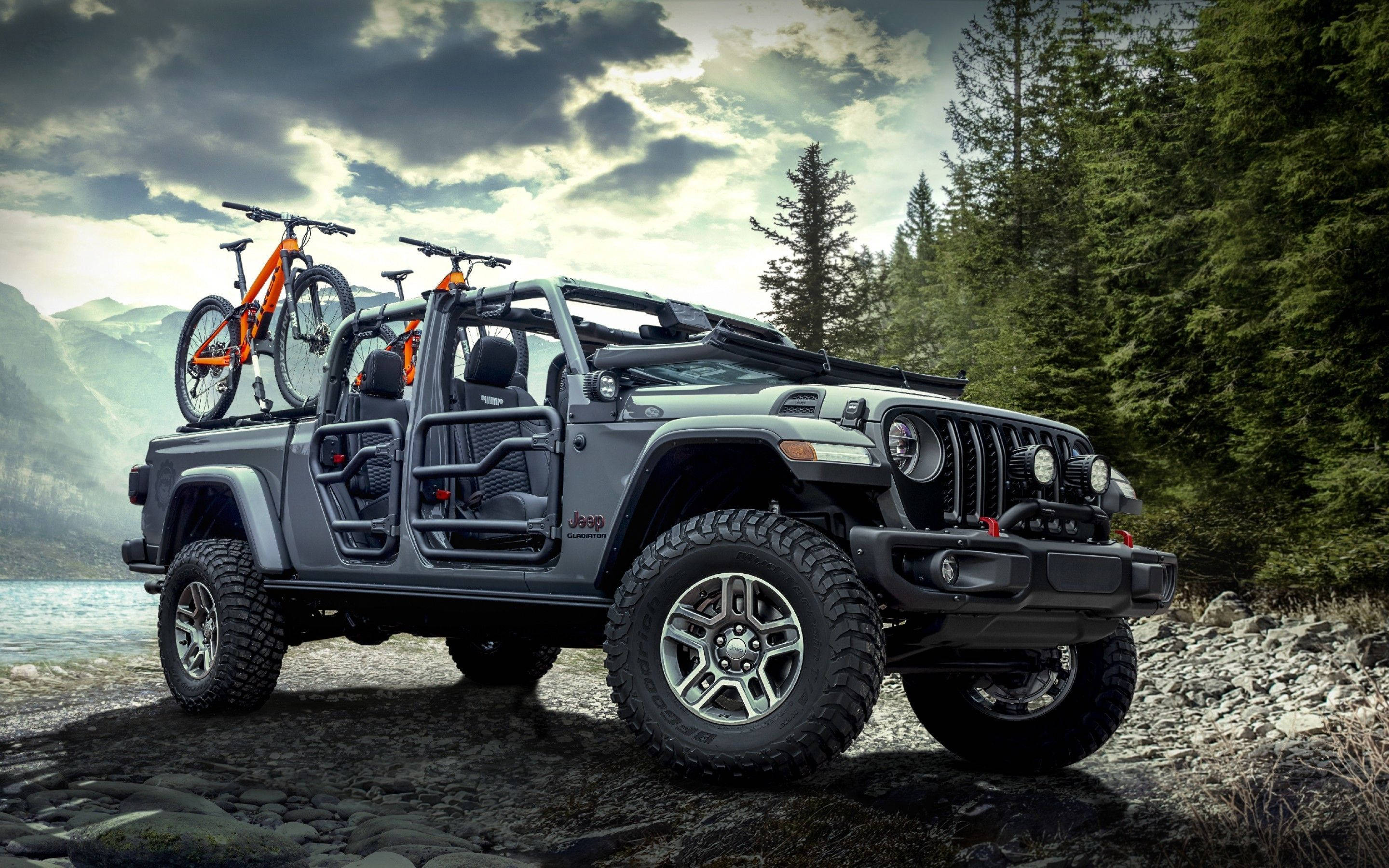 吉普 桌布 壁紙 — 一輛灰色Jeep Wrangler車頂綁著兩輛橙色登山自行車，停在岩石湖岸上，天空多雲而戲劇化。