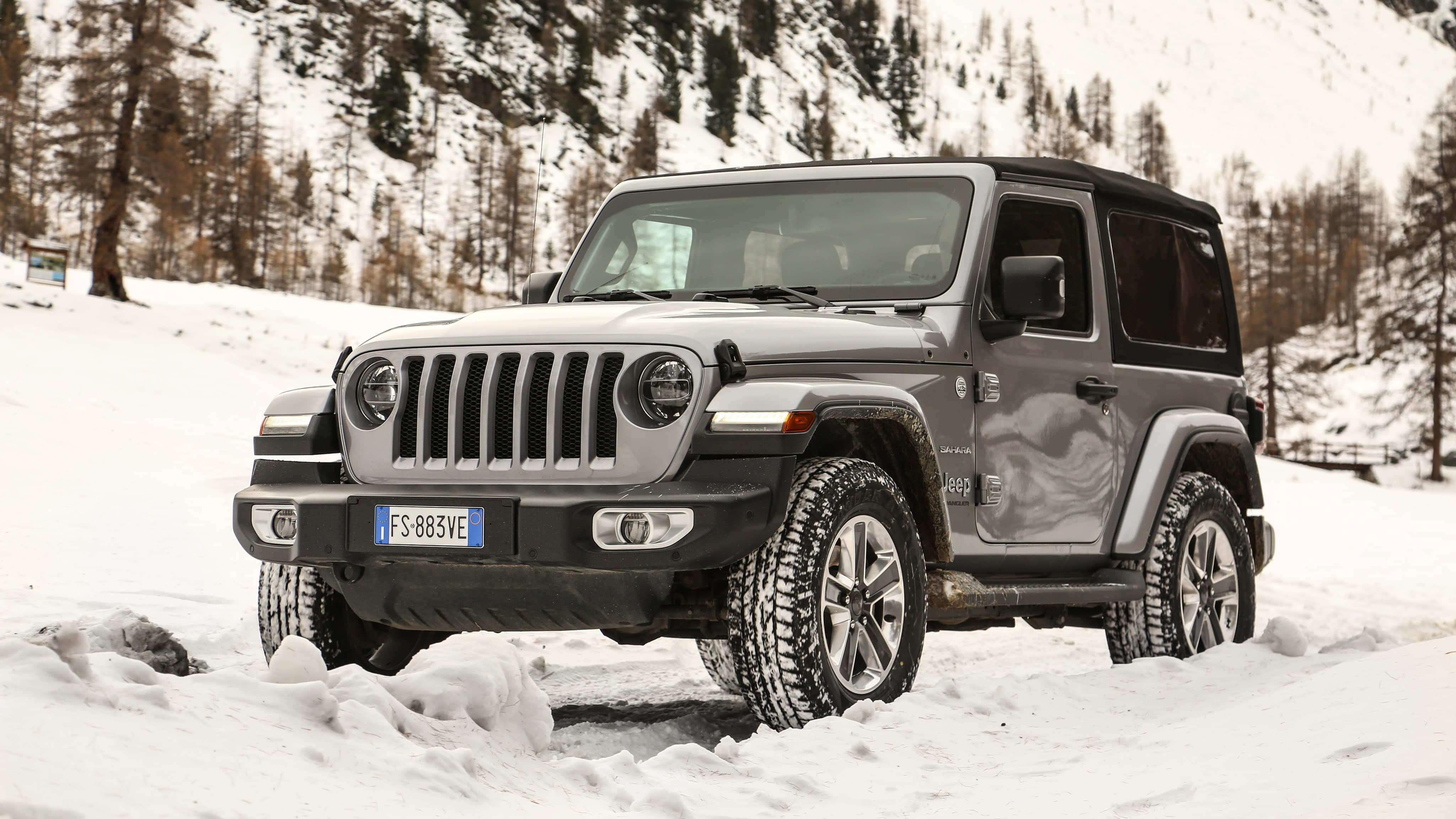 Jeep Wrangler 桌布 壁紙 — 一輛灰色Jeep Wrangler在積雪深厚的雪地中行駛，附近有被雪覆蓋的松樹和岩石山腰。