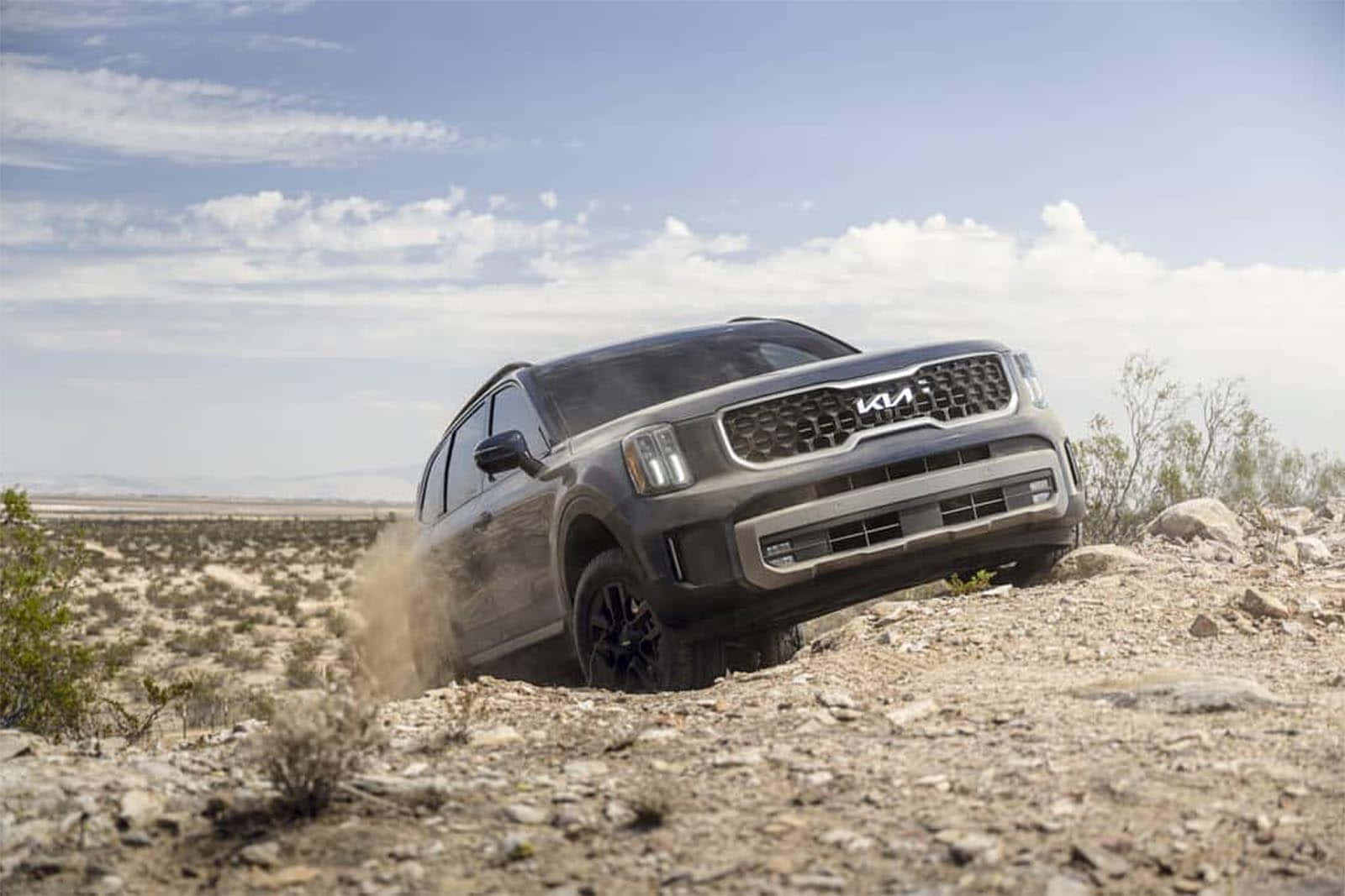 Kia Telluride 桌布 壁紙 — 一輛灰色Kia SUV在岩石沙漠地形上行駛，在零星雲朵的明亮藍天下揚起灰塵。