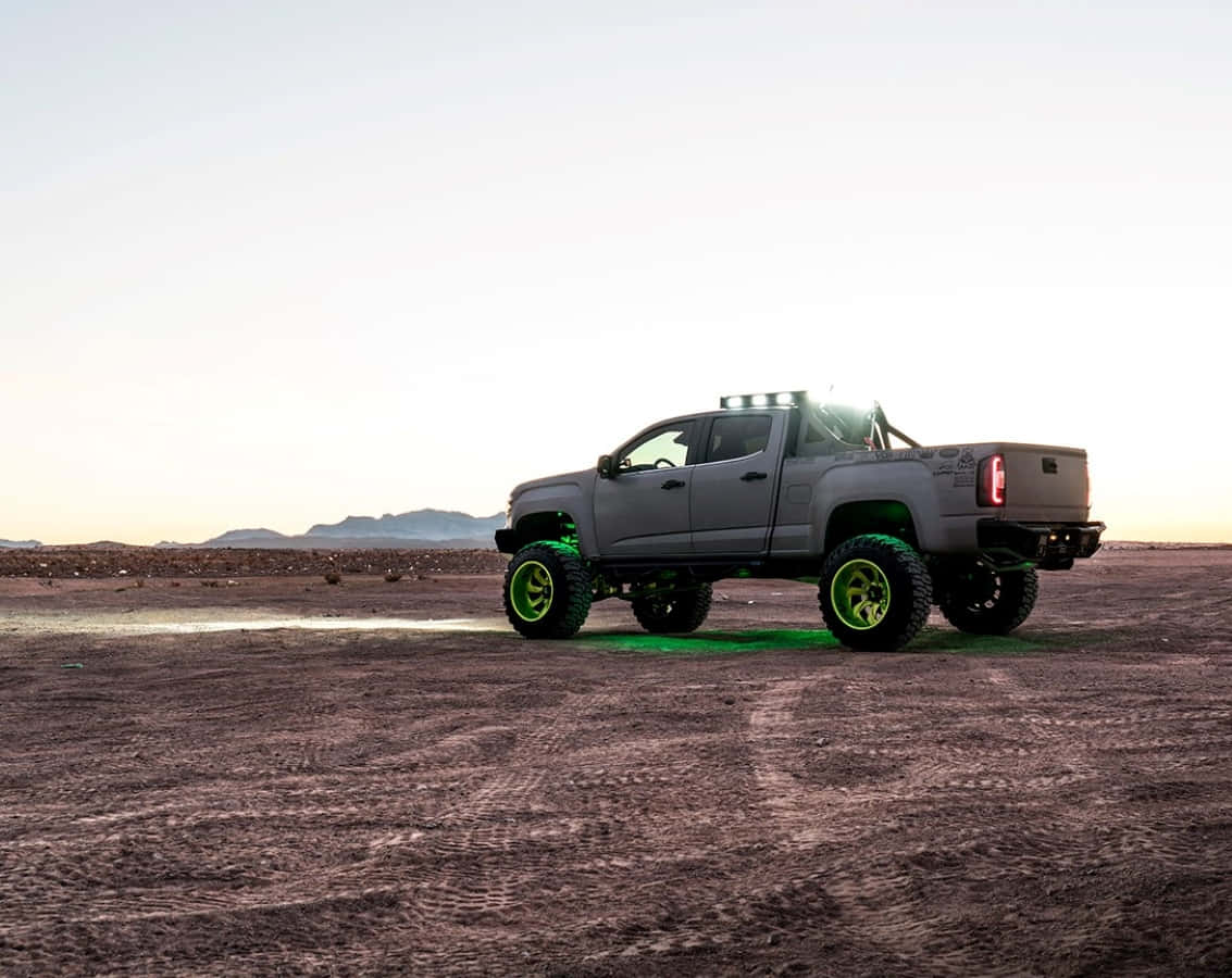 Gmc Canyon 桌布 壁紙 — 一輛灰色升高的皮卡車，帶有發光綠色車輪，站在廣闊的沙漠中，在日落時分於沙地上投下長長的影子。