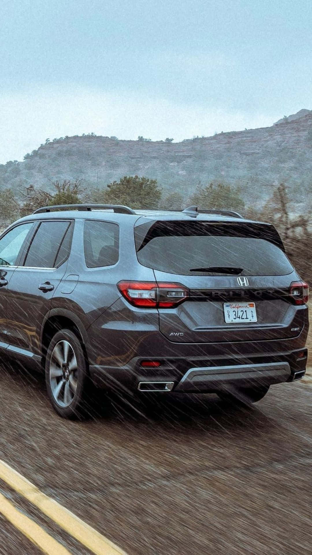 2023 Honda Pilot 桌布 壁紙 — 一輛灰色本田Pilot SUV在暴雨中行駛於山路，背景有模糊的動態和霧氣籠罩的山丘。