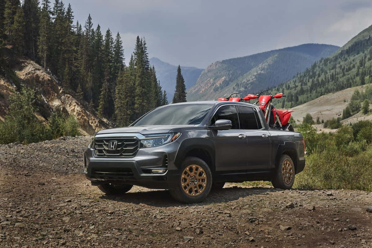 Honda Ridgeline 桌布 壁紙 — 一輛灰色本田Ridgeline皮卡車，帶有金色輪圈，車斗上載著一輛紅色摩托車，停泊在岩石山徑上，周圍是常綠森林和遠處的山丘，天空多雲。