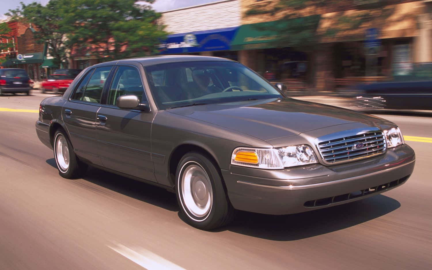 Ford Crown Victoria 桌布 壁紙 — 一輛灰色福特皇冠維多利亞轎車在陽光明媚的城市街道上奔馳，背景的商店櫥窗呈現動態模糊效果。