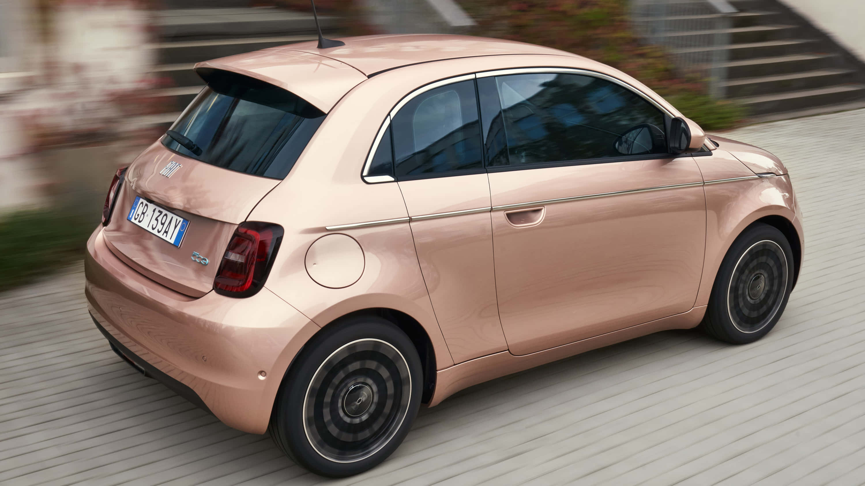 飛雅特 桌布 壁紙 — 一輛玫瑰金色的Fiat 500e電動車在模糊的城市環境中平穩地行駛在木質步道上。