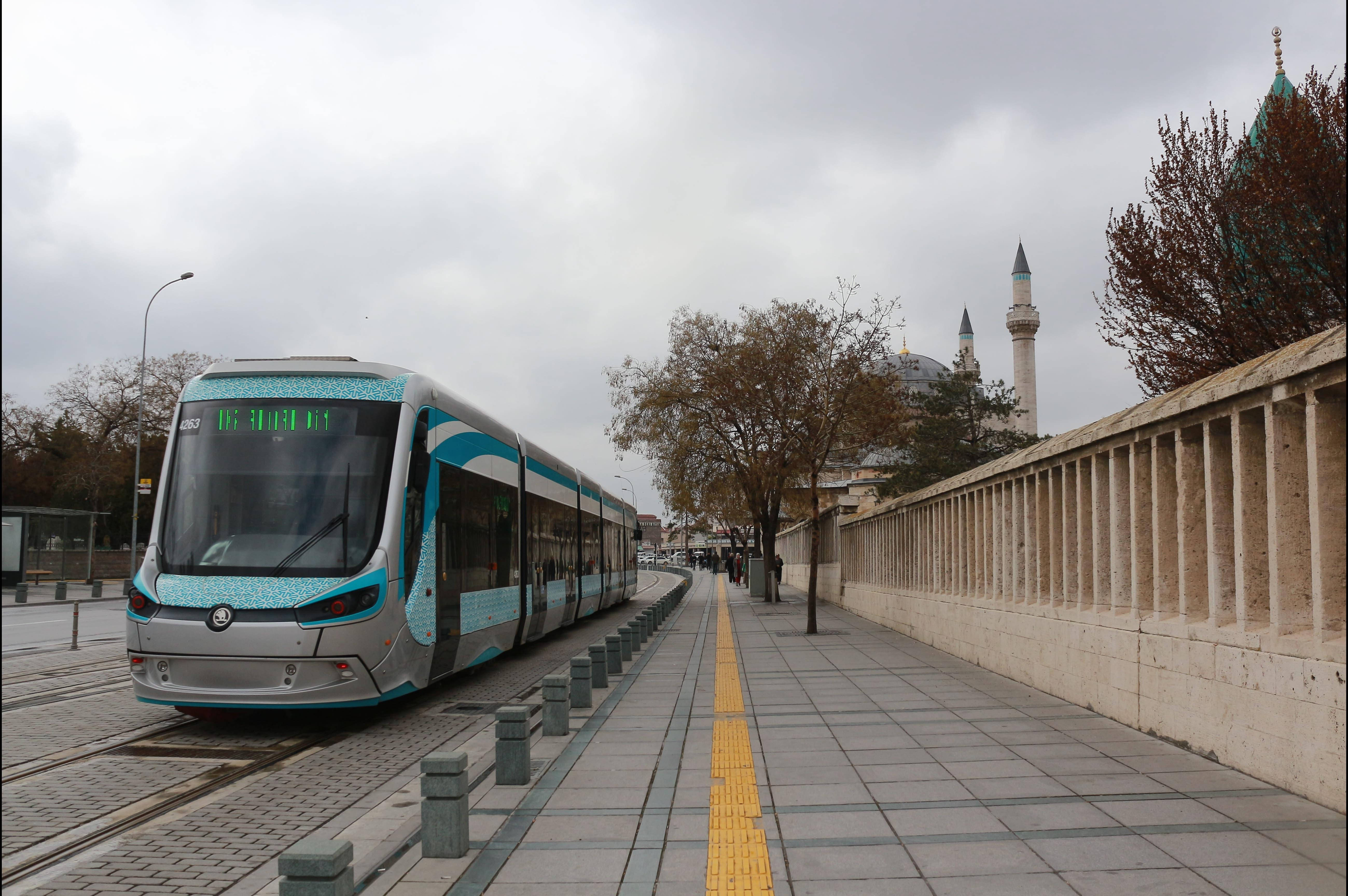 Konya 桌布 壁紙 — 一輛現代電車沿著鋪砌人行道旁的軌道滑行，旁邊有清真寺和秋天的樹木，天空多雲。