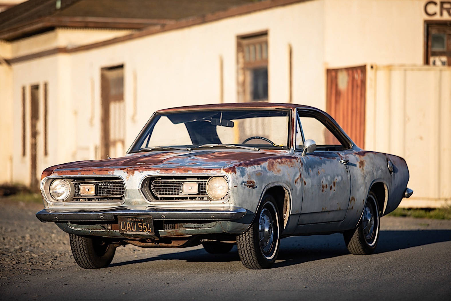 Barracuda 桌布 壁紙 — 一輛生鏽的復古汽車，油漆剝落，被遺棄在碎石地上，前面是一座風化的建築物，在溫暖的陽光下。