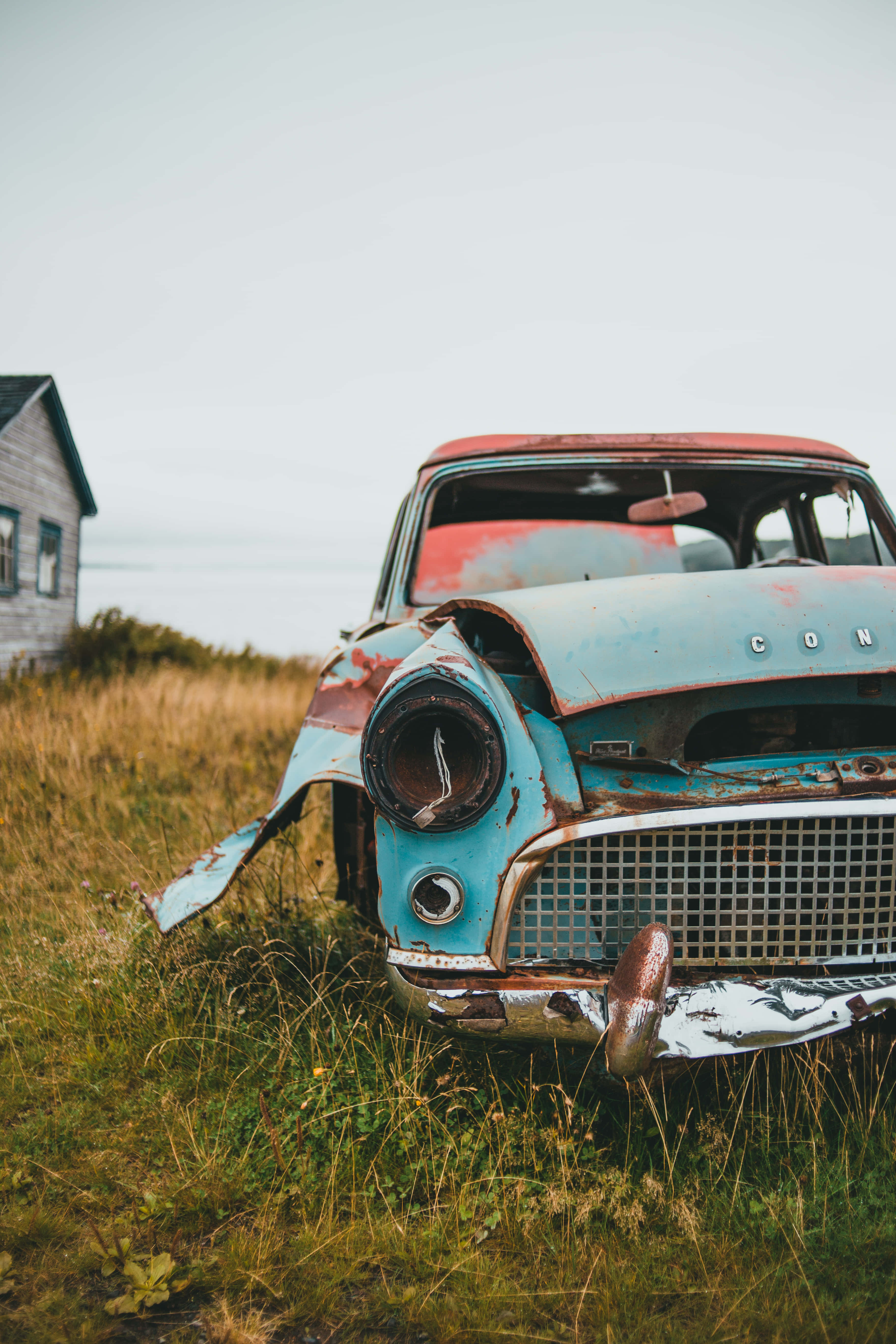 Vintage Car iPhone 桌布 壁紙 — 一輛生鏽的復古汽車，藍色和紅色油漆剝落，被遺棄在風化房屋附近的高草叢中。