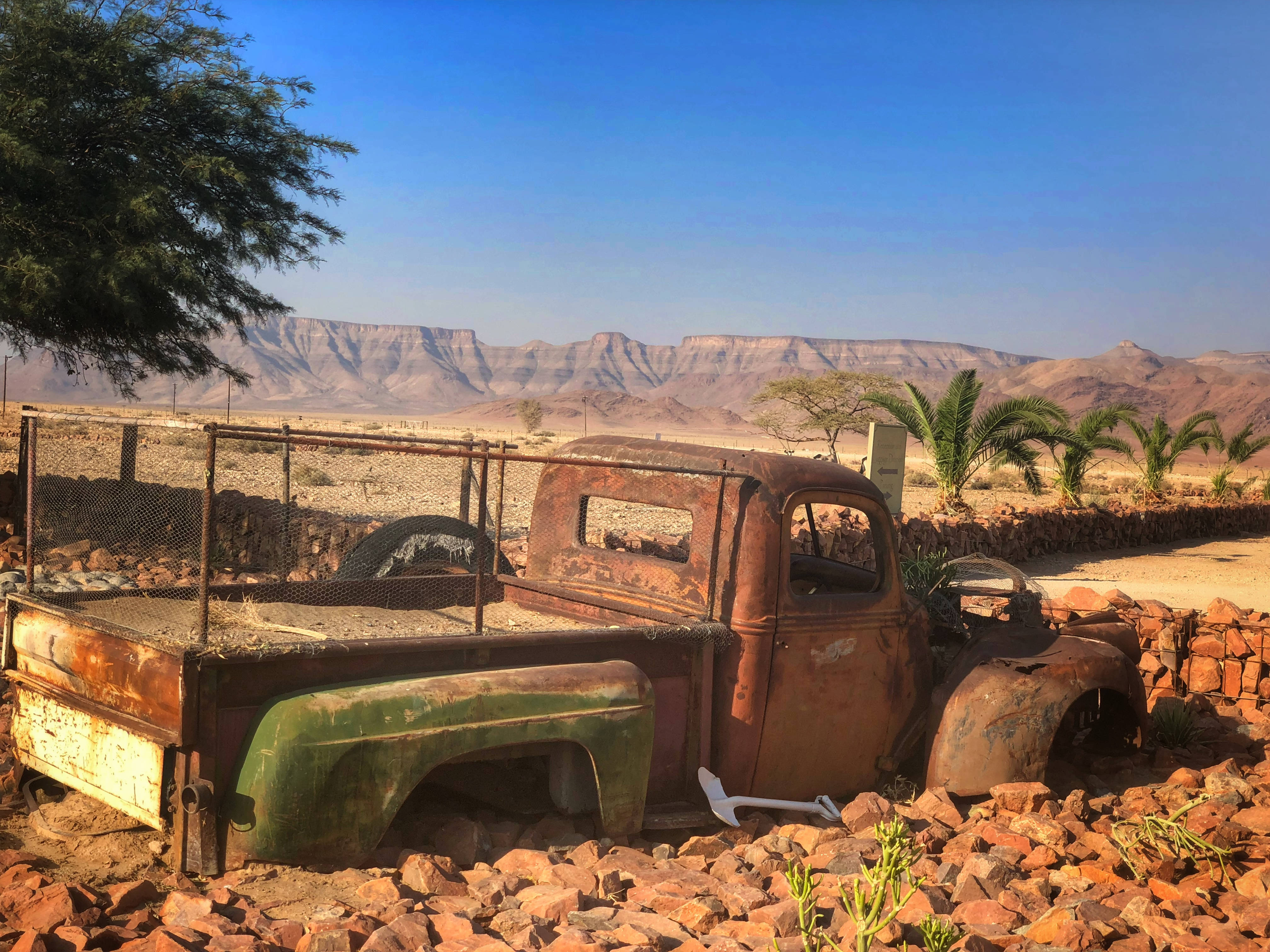 Namibia 桌布 壁紙 — 一輛生鏽的復古皮卡車被遺棄在紅色岩石間，背景是晴朗的藍天和遠處的沙漠山脈。