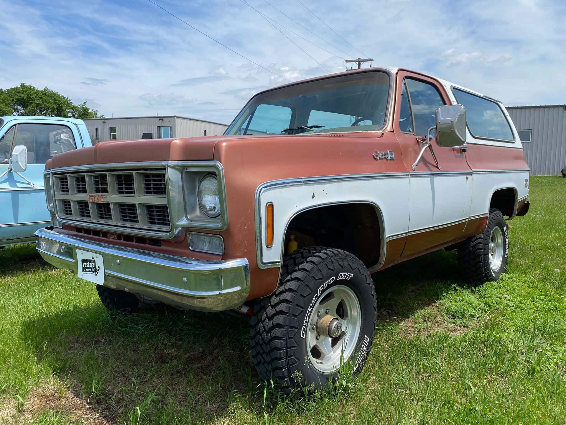 Gmc Jimmy 桌布 壁紙 — 一輛生鏽的橙色和白色復古GMC皮卡車，配有大尺寸輪胎，停在草地上，天空部分多雲。
