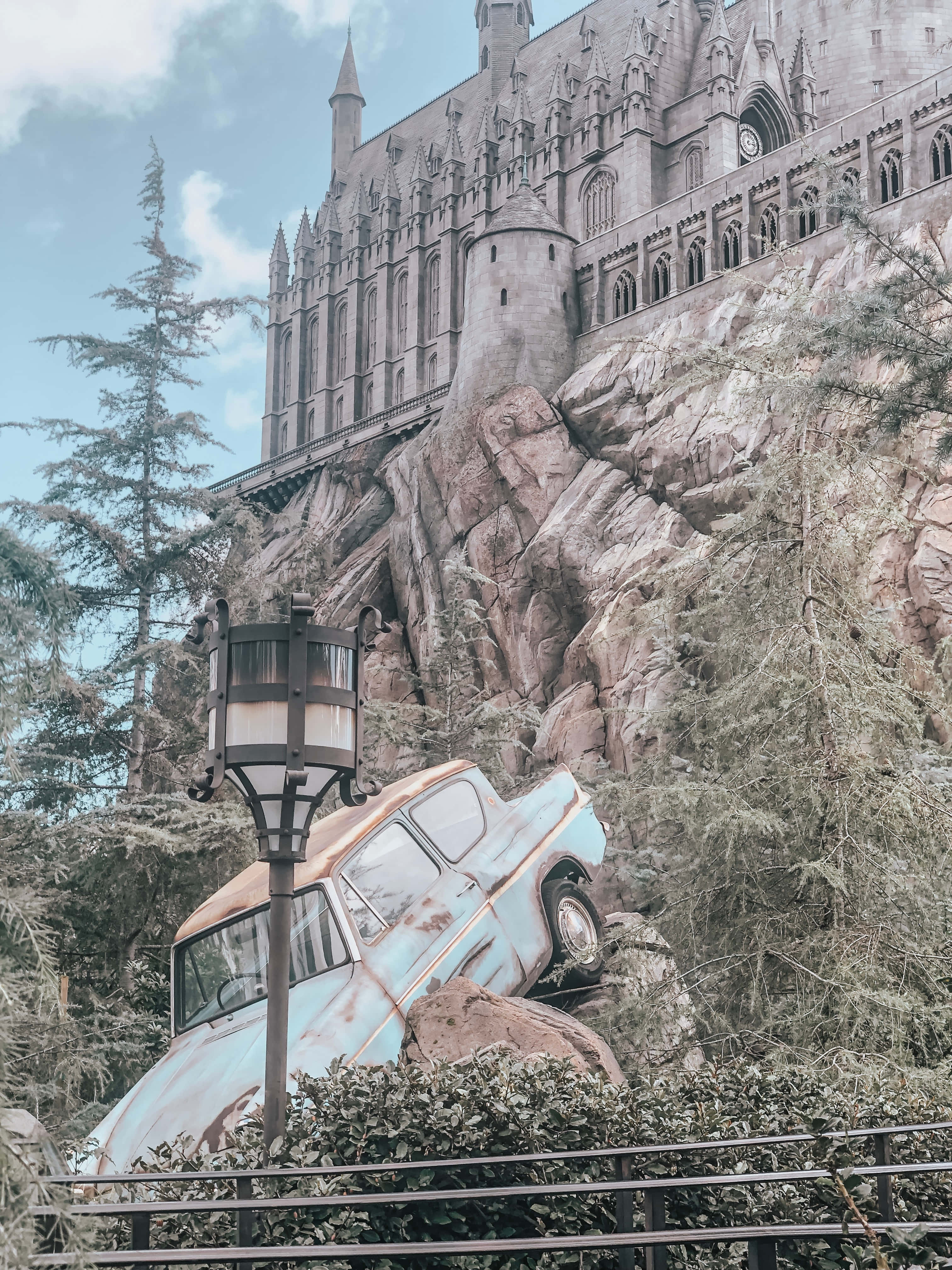 Wizarding World 桌布 壁紙 — 一輛生鏽的藍色汽車停放在岩石山坡上，下方是聳立的哥德式城堡，周圍環繞著常青樹。