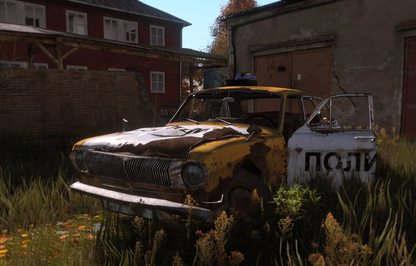DayZ 桌布 壁紙 — 一輛生鏽的黃色警車被遺棄在雜草叢生的廢棄建築旁，在明亮的日光下。