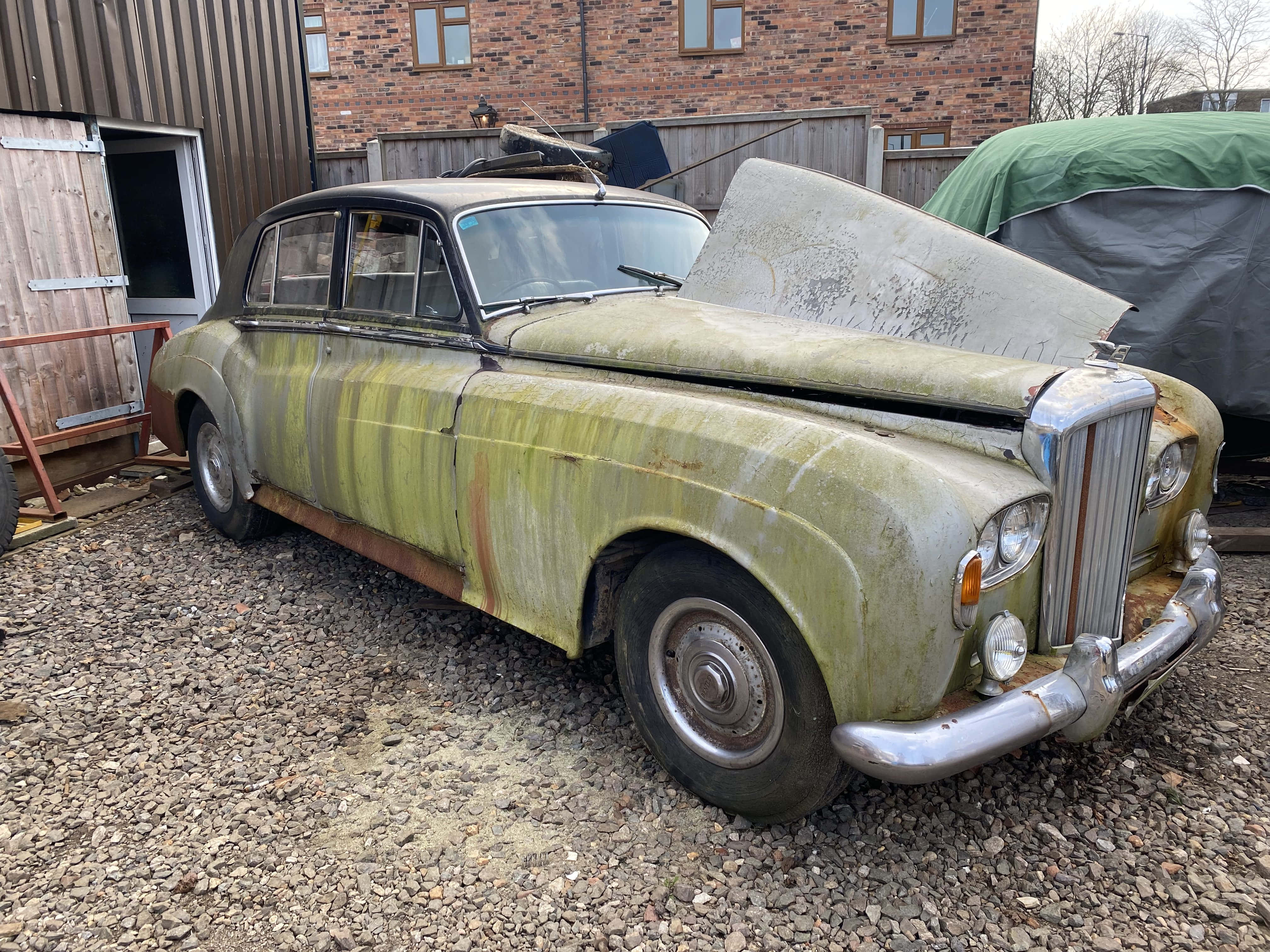 Bentley S3 桌布 壁紙 — 一輛生鏽長滿青苔的復古綠色汽車，引擎蓋打開，被遺棄在磚砌建築附近的礫石上。