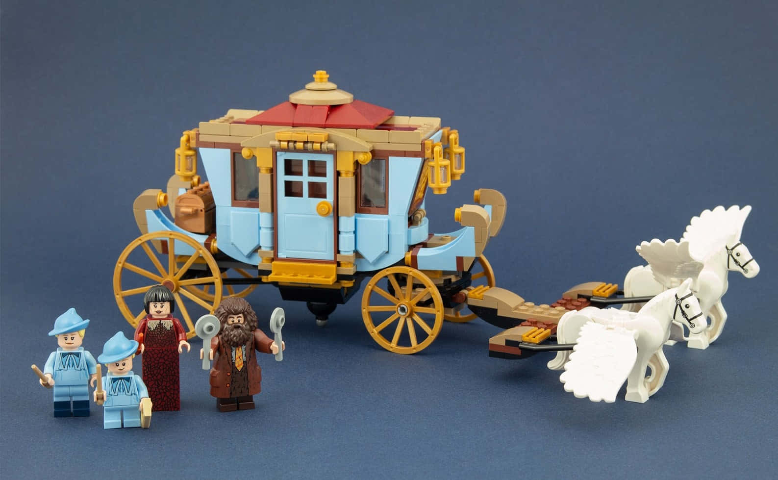 The Hogwarts Carriage 桌布 壁紙 — 一輛由兩匹白馬拉動的LEGO西部驛馬車，旁邊站著四個迷你人偶，背景為純藍色。