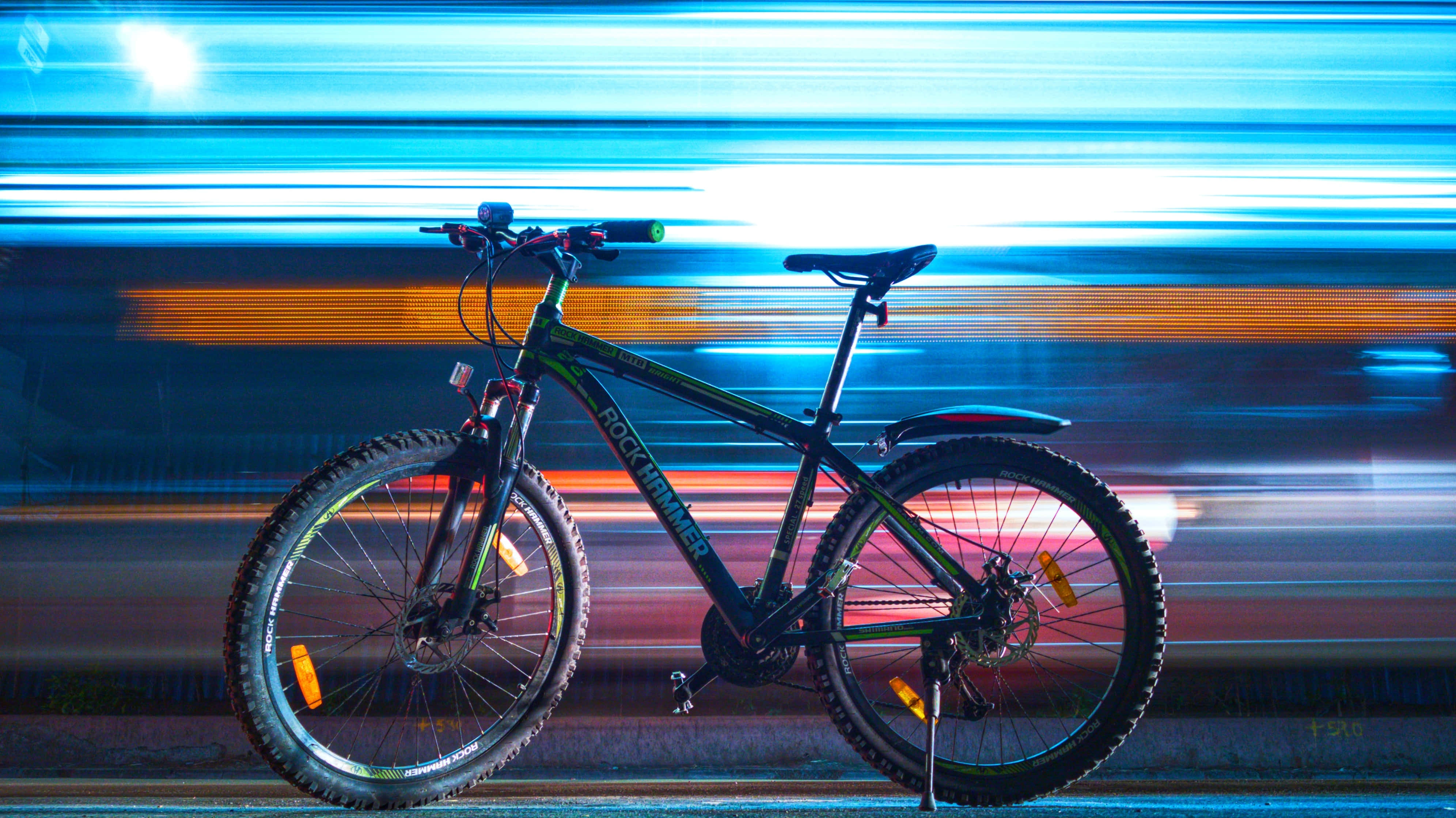 Mountain Bike 桌布 壁紙 — 一輛登山車在夜晚的霓虹城市景觀前靜止，帶有藍色和橙色的光跡。
