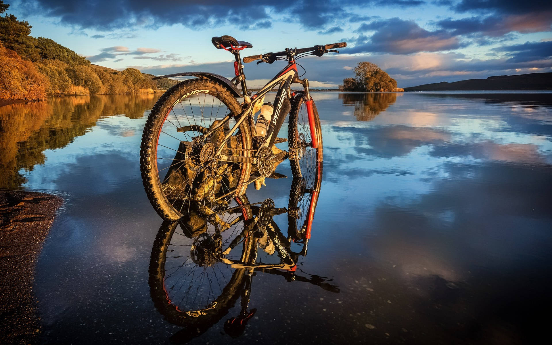 Mountain Bike 桌布 壁紙 — 一輛登山車站在淺水中，倒映著秋樹環繞的平靜湖面上戲劇性的日落天空。