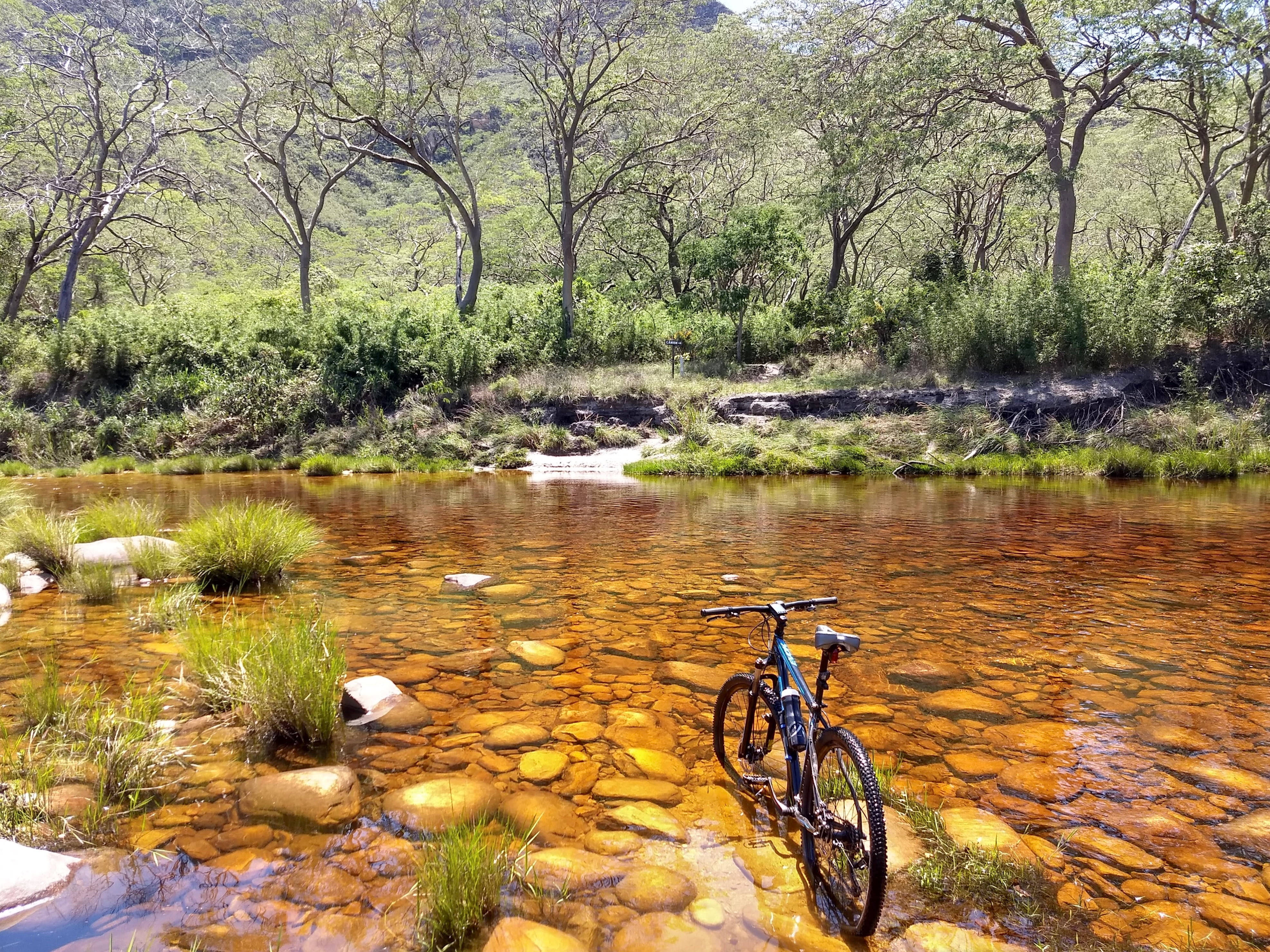 Mountain Bike 桌布 壁紙 — 一輛登山車站在淺琥珀色水中，下方是光滑的石頭，周圍是茂密的綠樹和山丘，在明亮的陽光下。