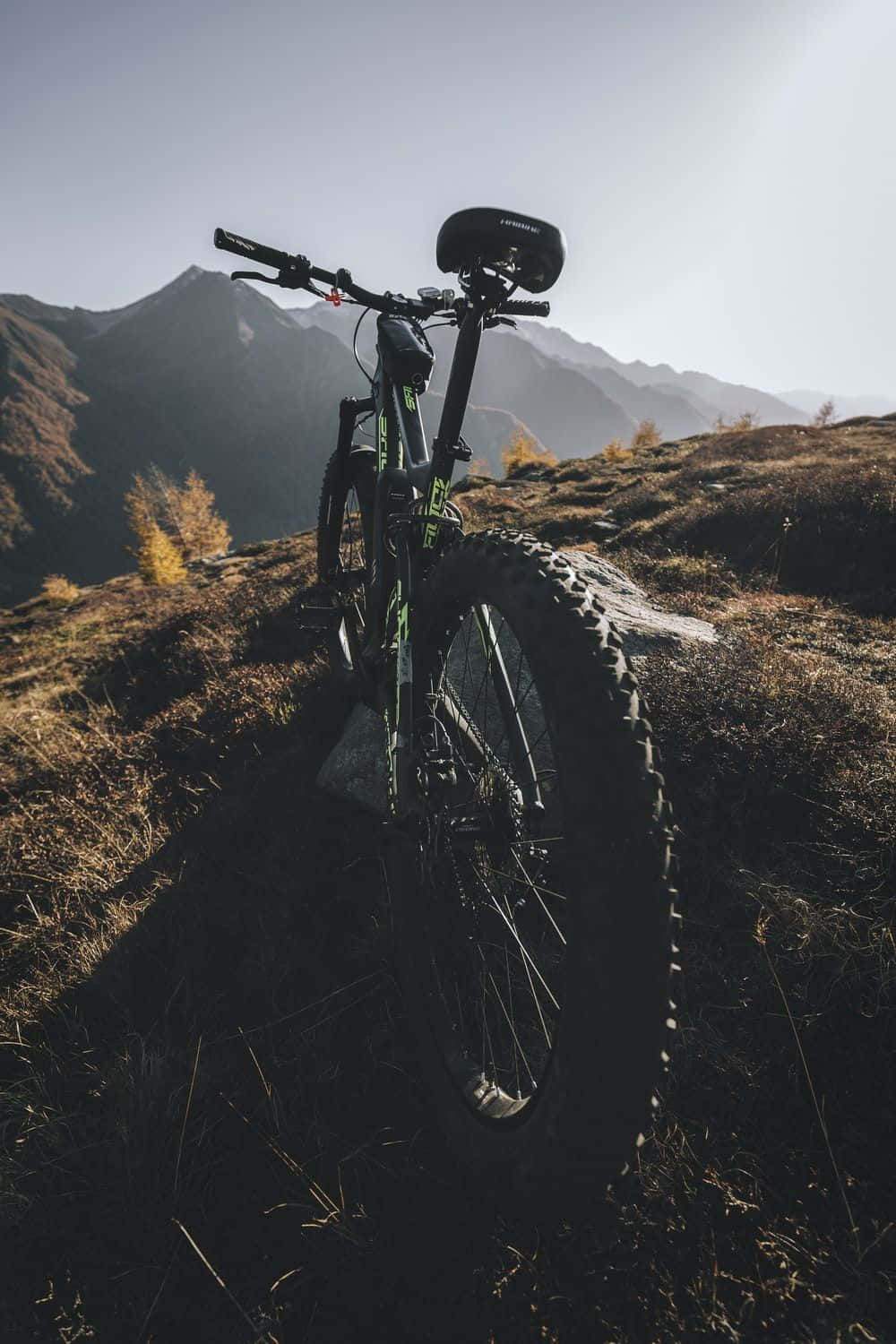 Mountain Bike 桌布 壁紙 — 一輛登山車豎立在長滿草的山丘上，背景有崎嶇的山峰和金色的樹葉，天空明亮。