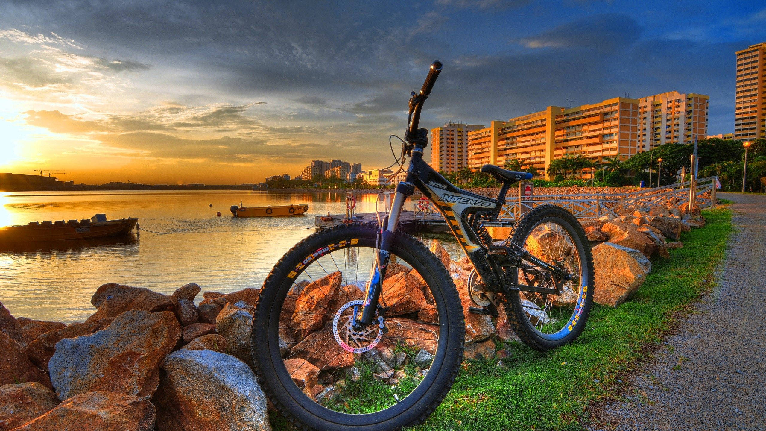 Mountain Bike 桌布 壁紙 — 一輛登山車靠在水邊小徑旁的石頭上，夕陽正落在城市天際線和寧靜的水面上。