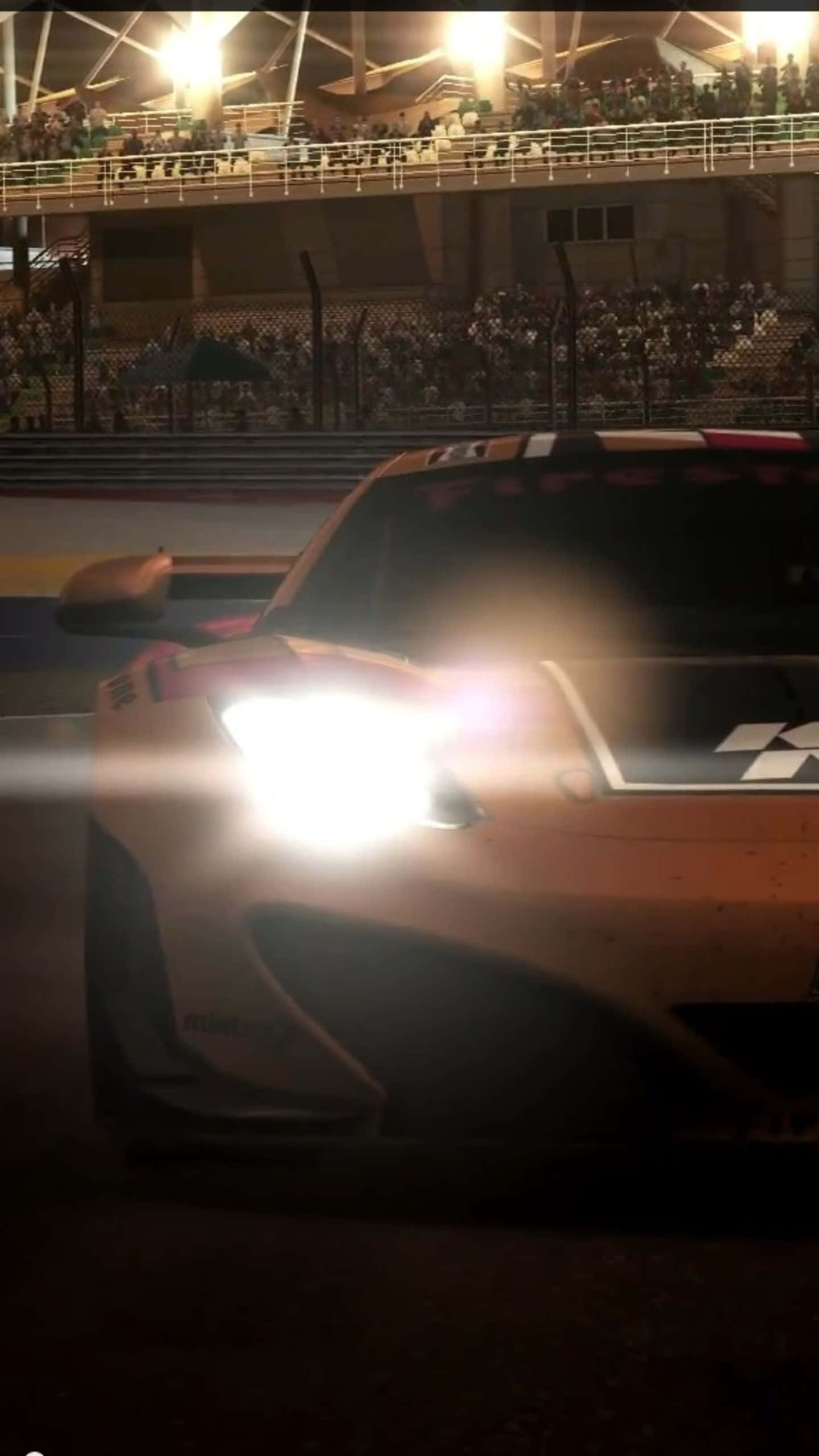 Android Grid Autosport 桌布 壁紙 — 一輛發光的橙色賽車的頭燈劃破夜空，在體育場賽道上飛馳而過，周圍是歡呼的人群。