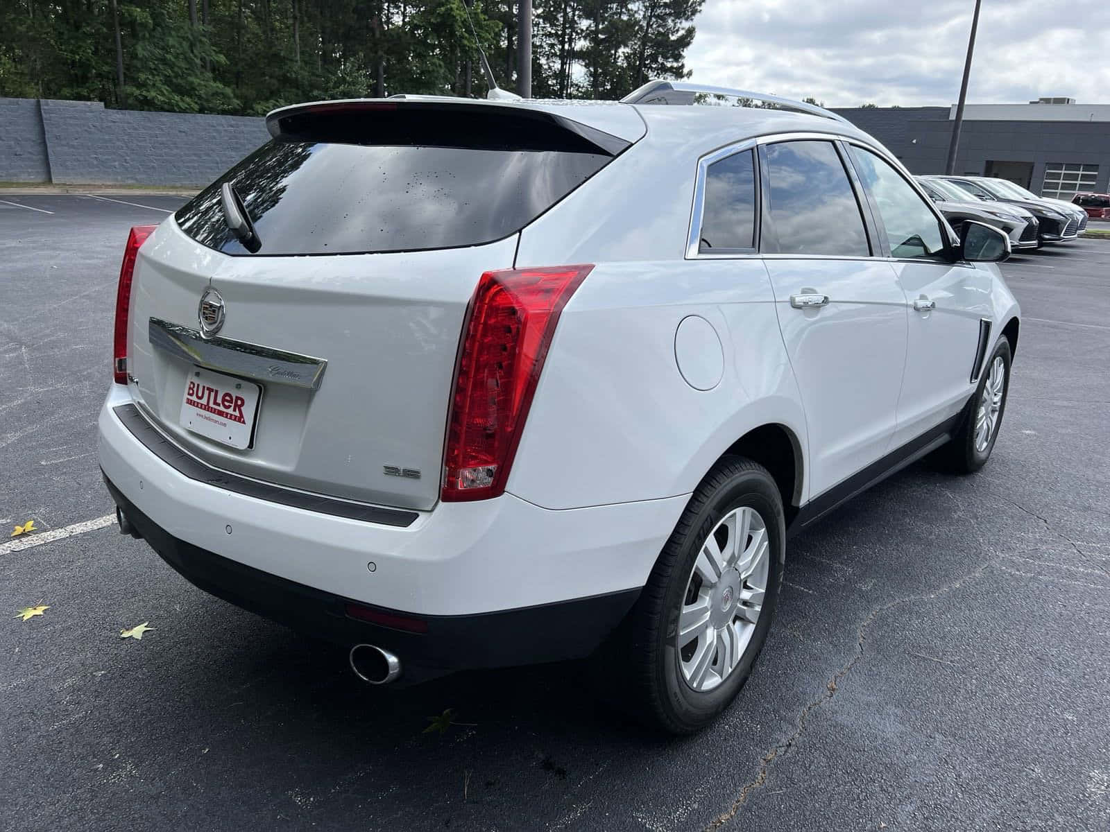 Cadillac Srx 桌布 壁紙 — 一輛白色 Cadillac SRX SUV 停放在瀝青停車場，背景有樹木和建築物，天空多雲。